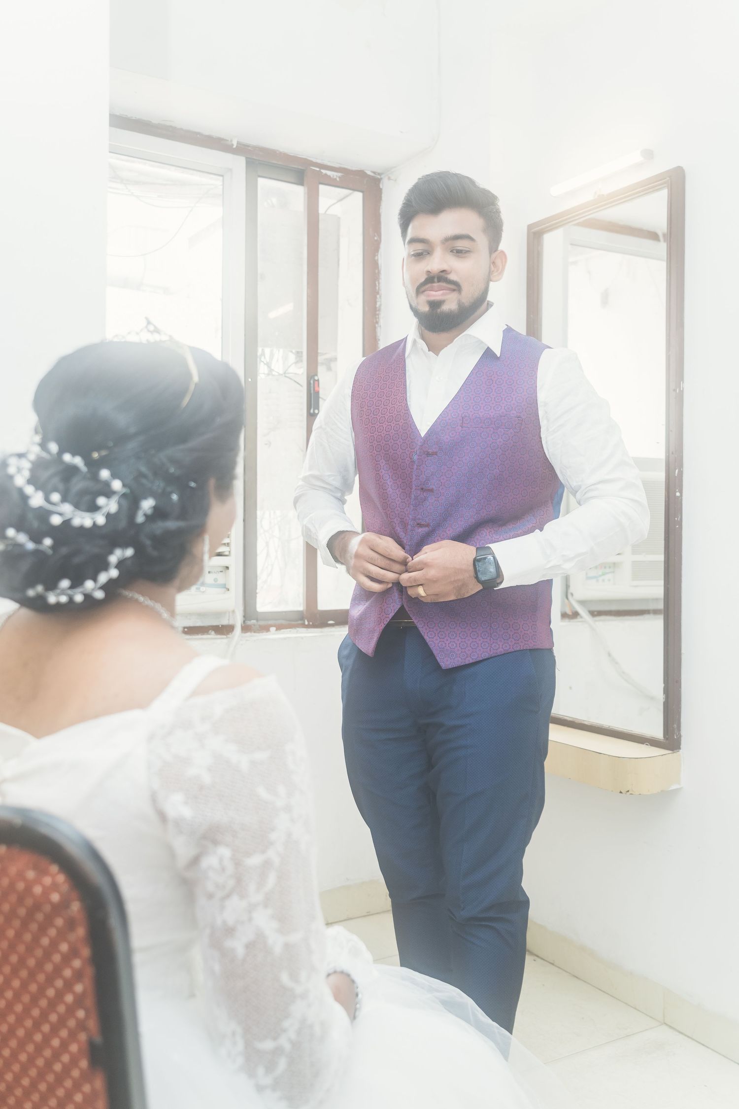 A couple in wedding attire sharing a moment in front of mirrors in a bright white room.