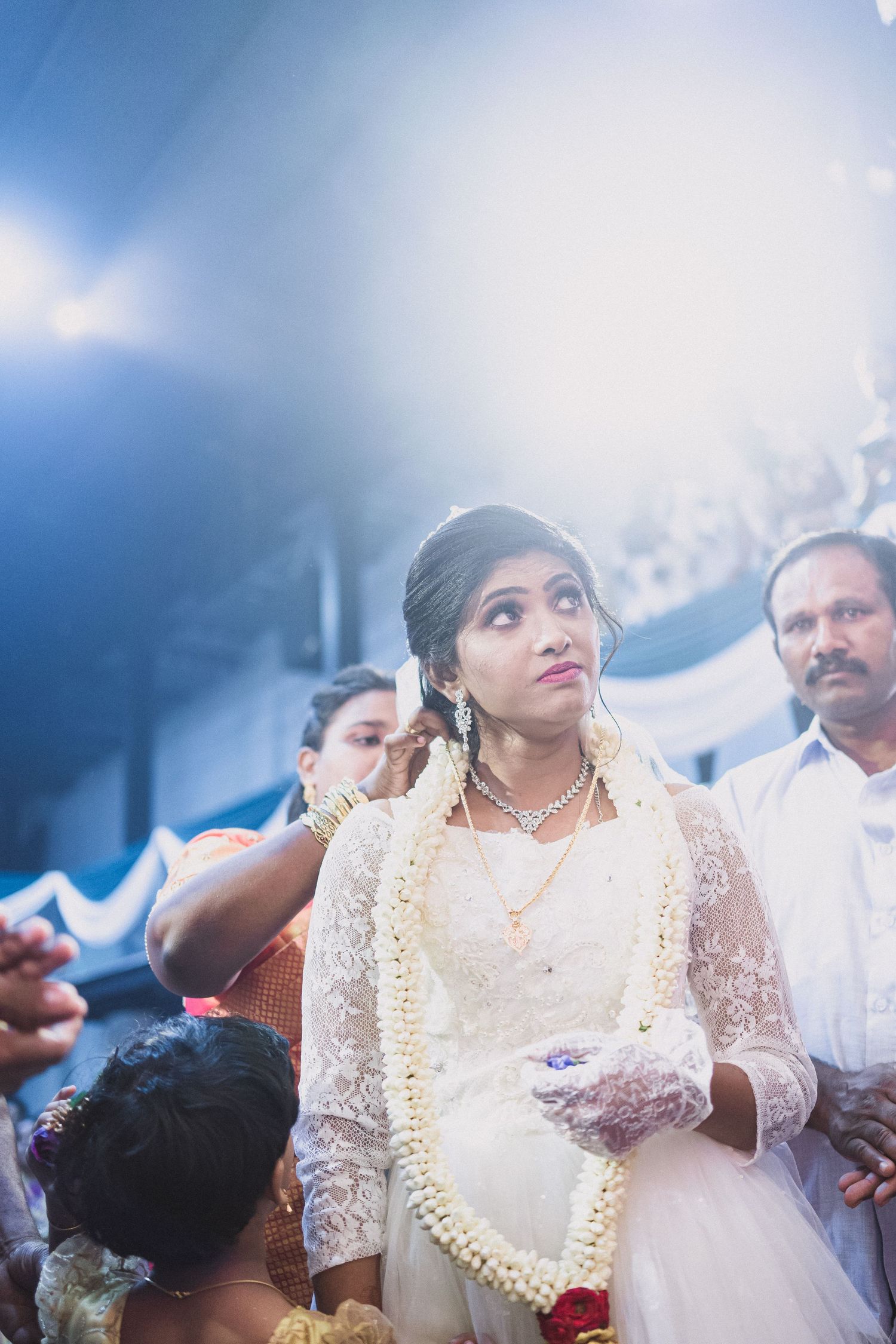 Bride in white lace dress and long jasmine garland looks up during traditional Christian wedding ceremony.