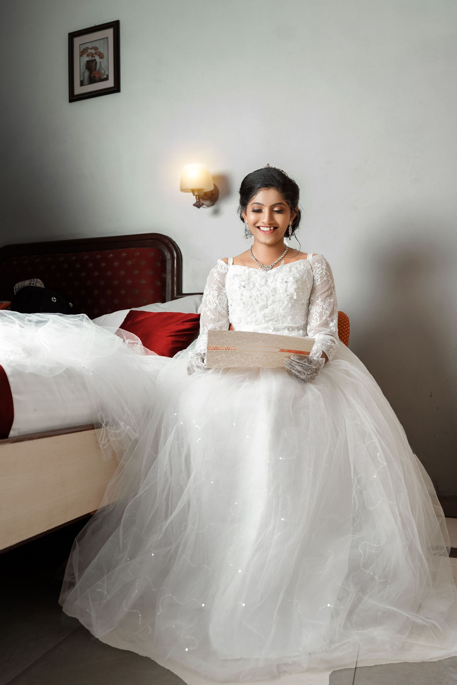 A radiant bride in a white wedding gown sits on a bed with a small frame on the wall.