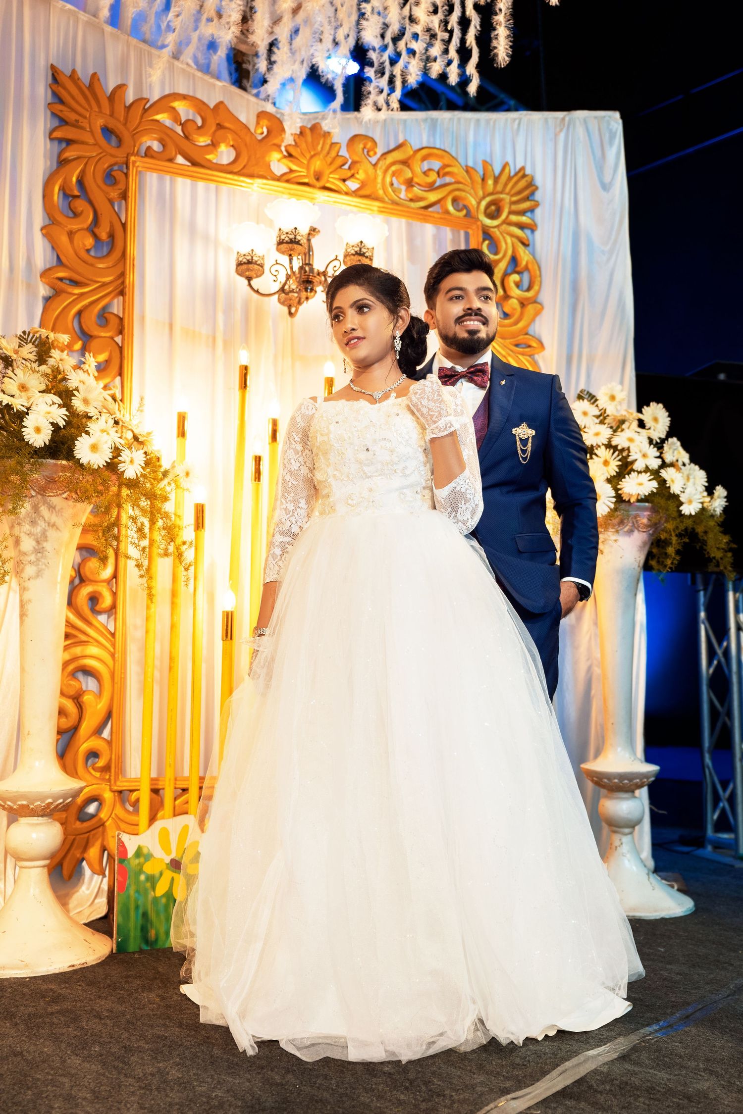 Bride and groom pose elegantly in front of an ornate golden frame and floral decorations.