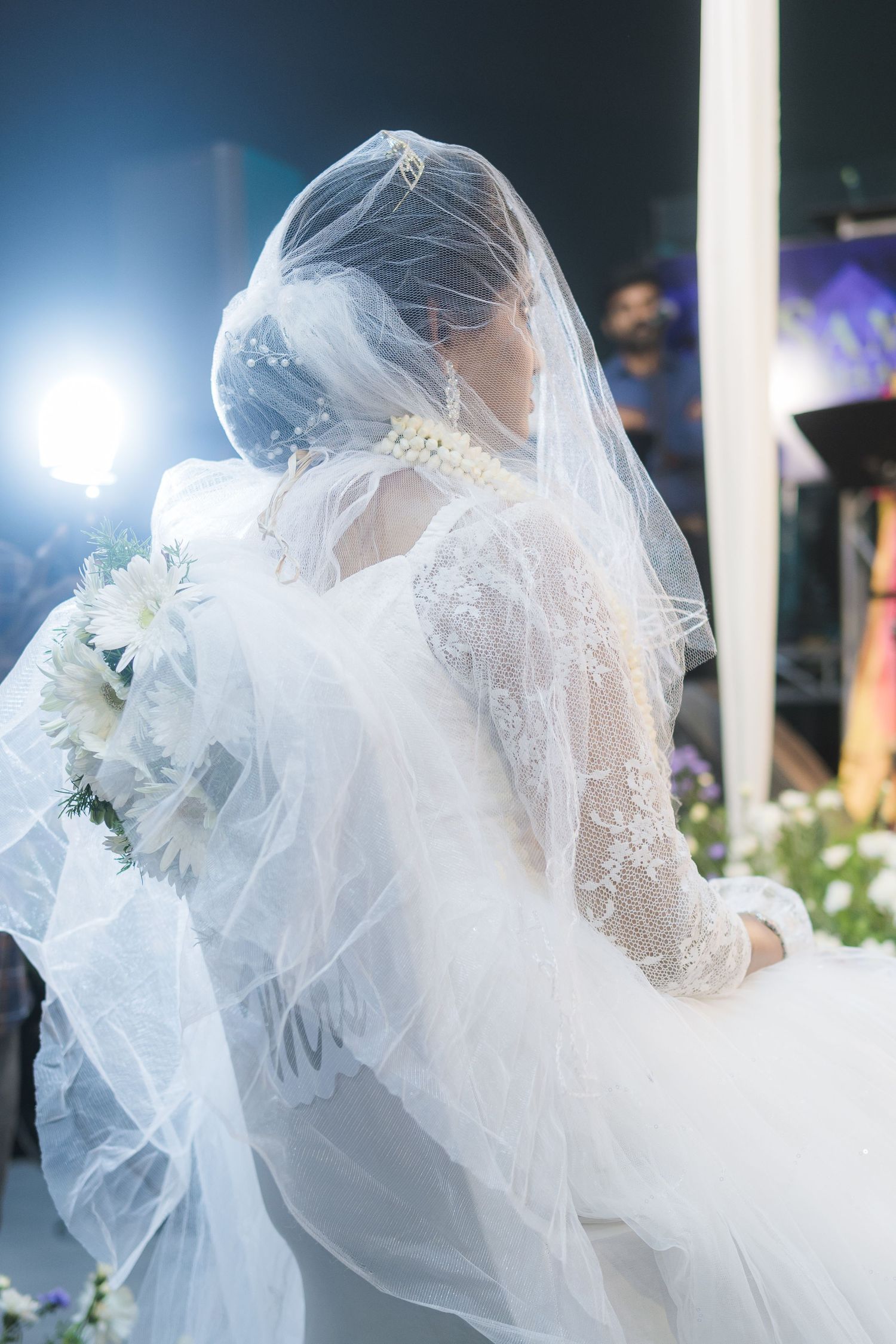 Bride wearing intricate lace wedding gown and flowing veil captured in dreamy backlit photograph.