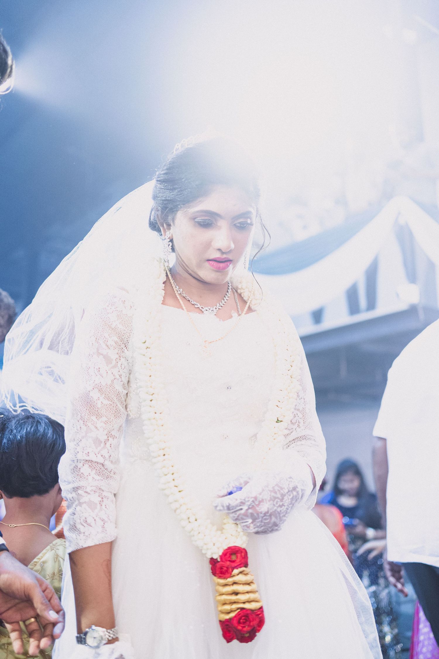 Bride wearing white gown and holding red flowers stands during traditional ceremony.