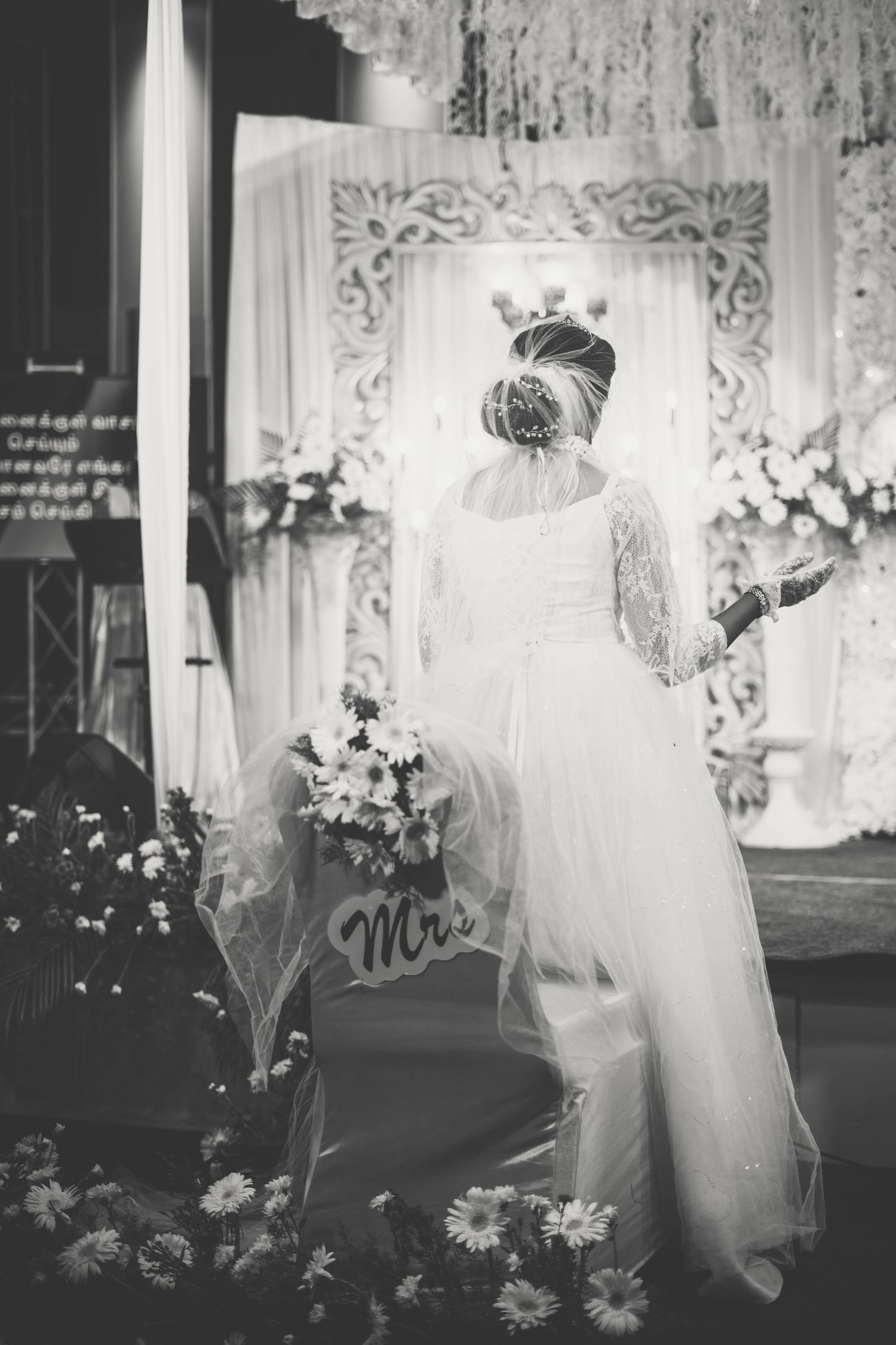 A bride in an elegant white lace gown and veil stands in front of an ornate backdrop at her wedding.