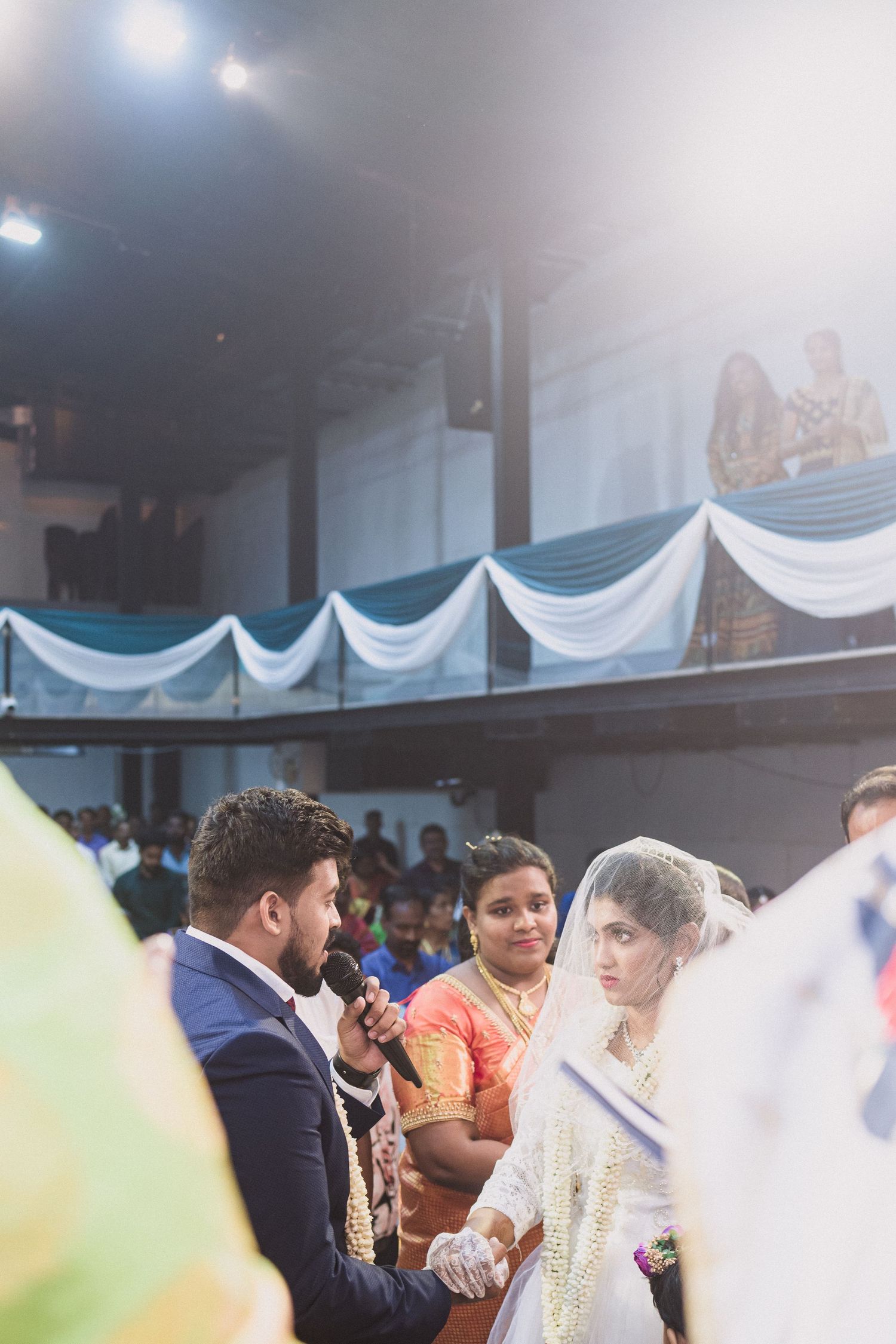 Wedding ceremony in a hall decorated with white drapes and bright overhead lighting.