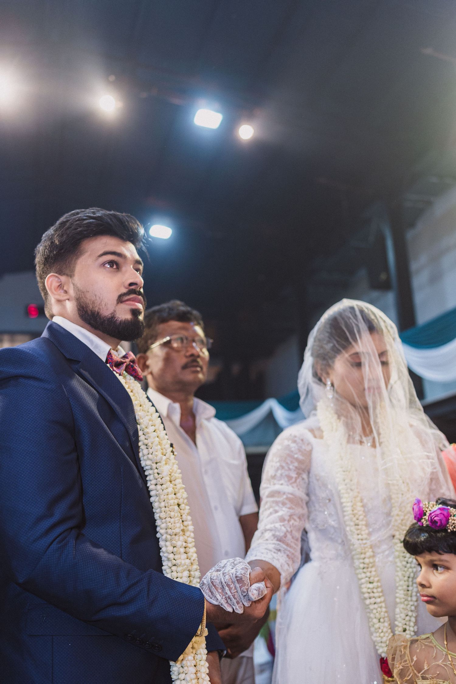 A wedding ceremony with traditional floral garlands and white decor highlights the bride in a veil and groom in navy suit.