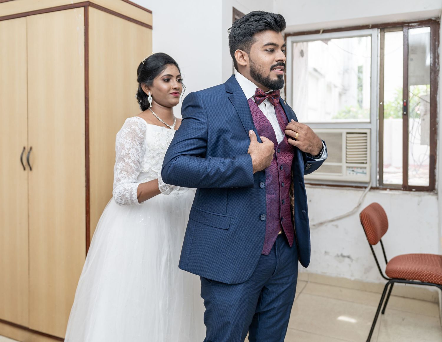 A couple helping each other with final wedding attire adjustments in a dressing room.