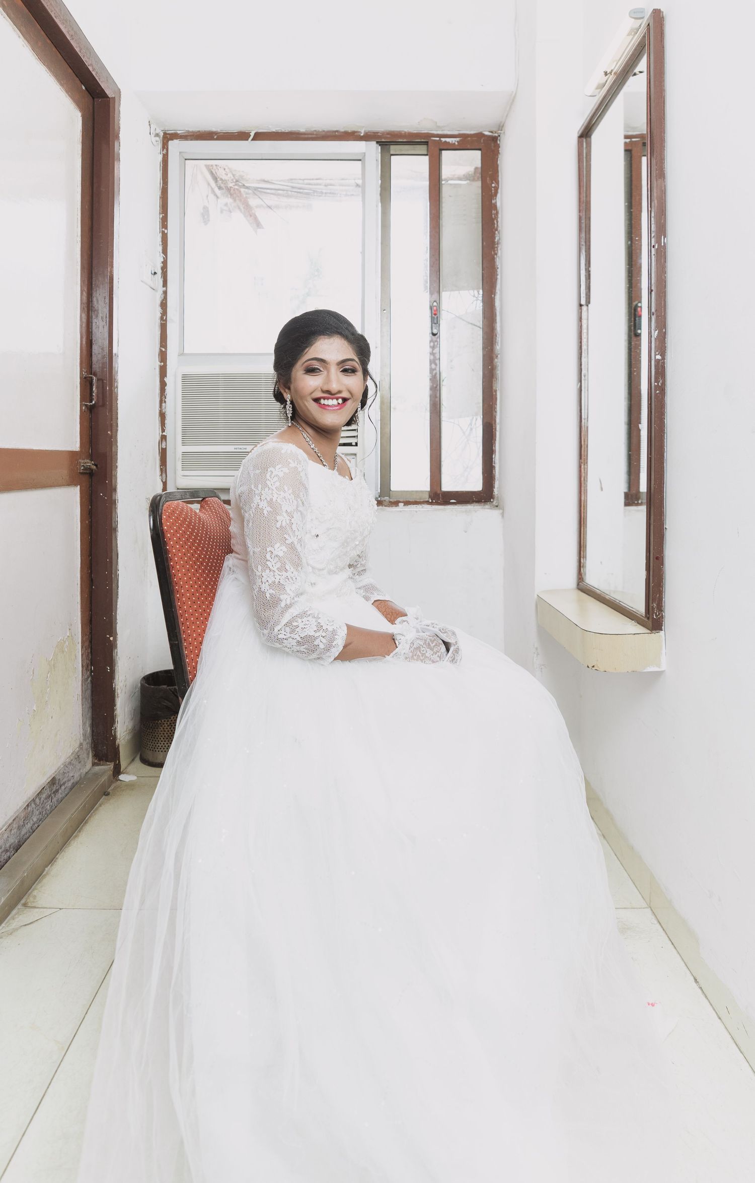 An elegant bride in a white lace gown sits in a chair in a bright hallway.