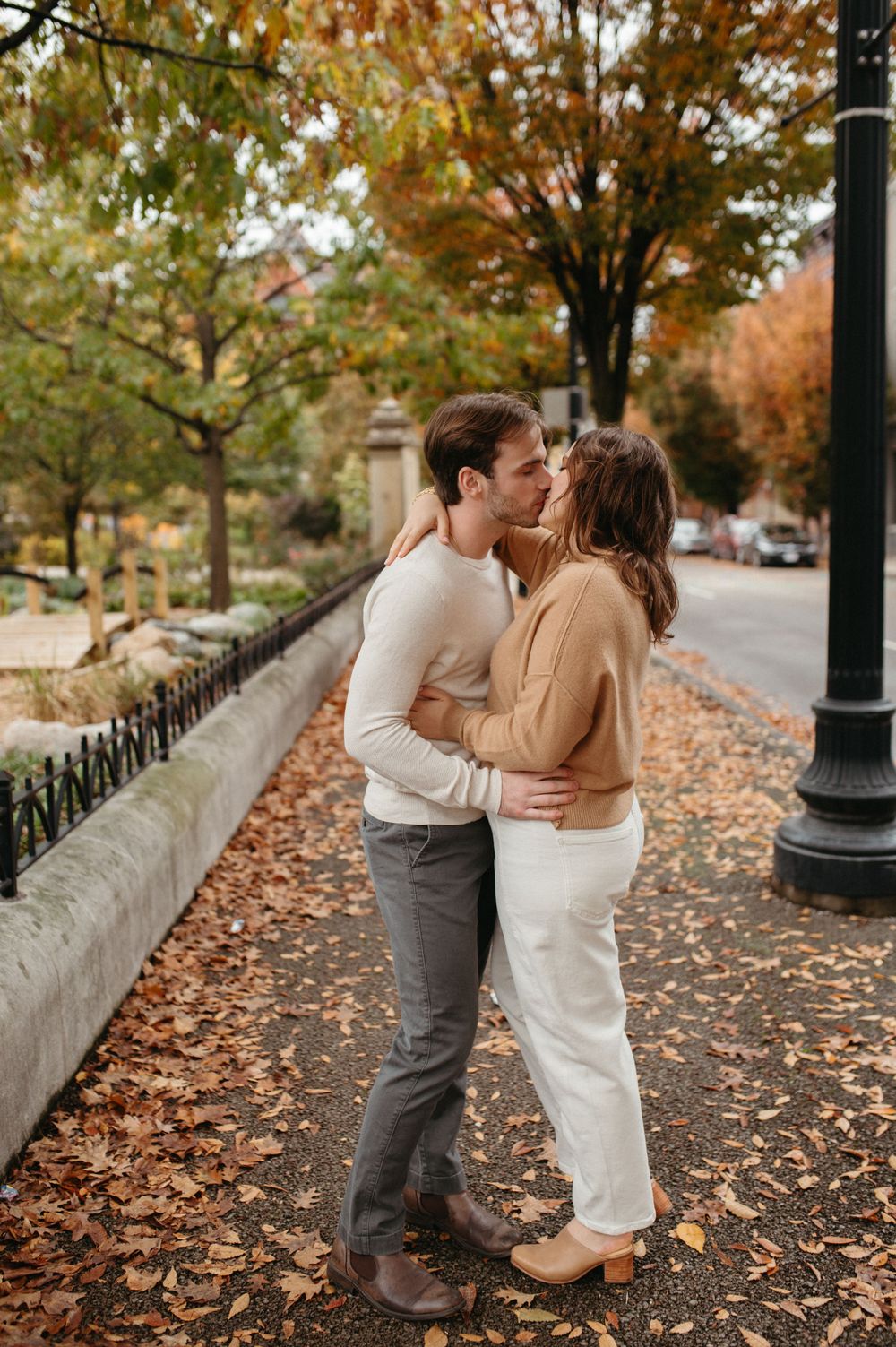 Romantic couple embraces on an autumn sidewalk lined with fallen leaves and trees.