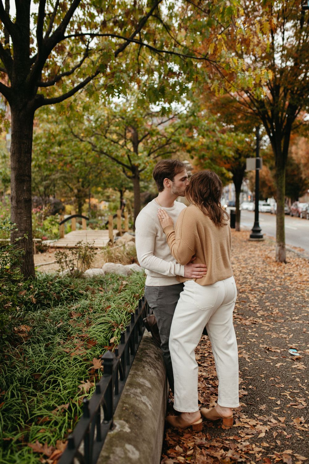 Couple sharing romantic moment by iron fence with autumn leaves scattered around.