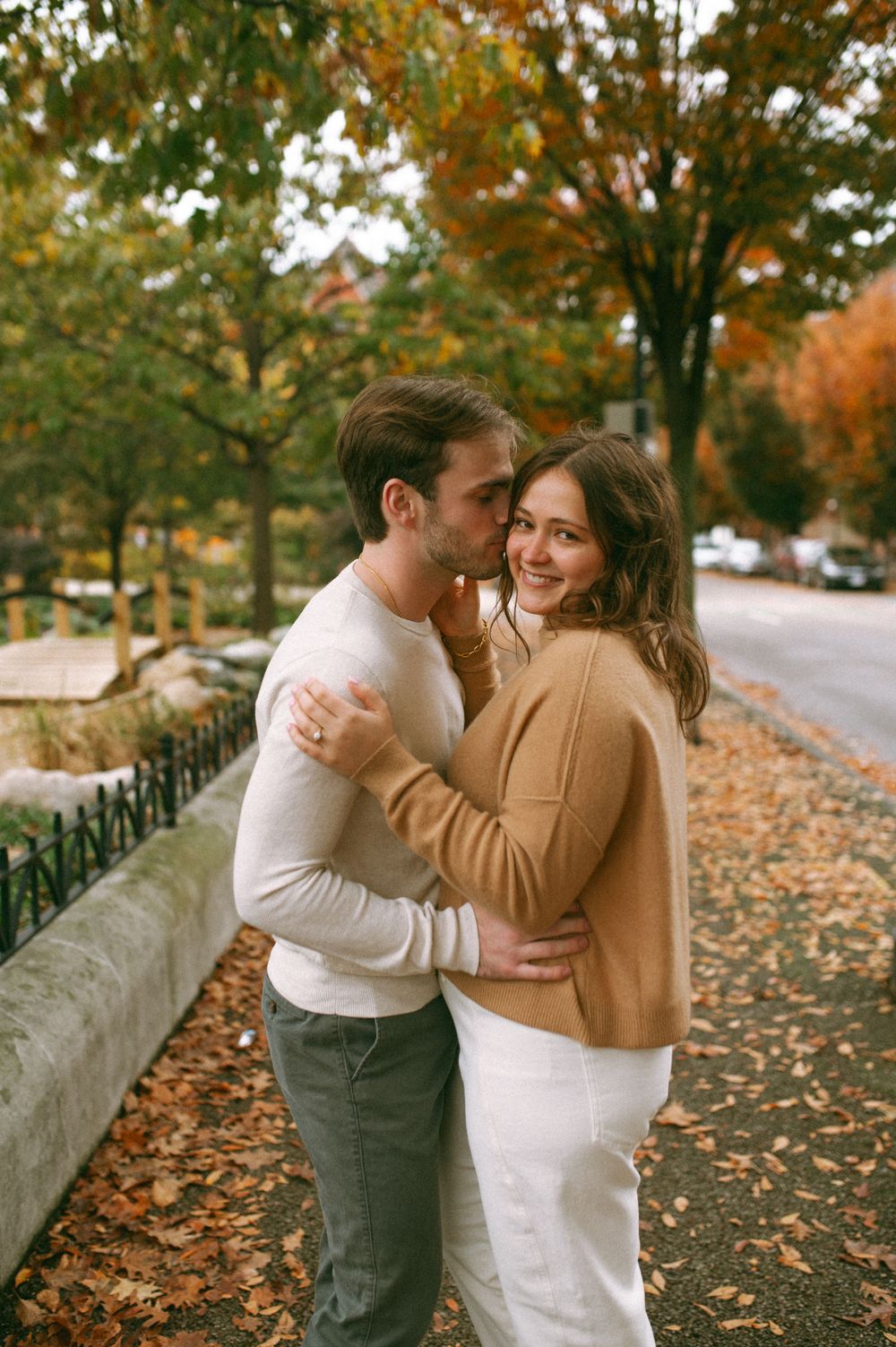 Couple in casual attire embracing on sidewalk with fall foliage in background.