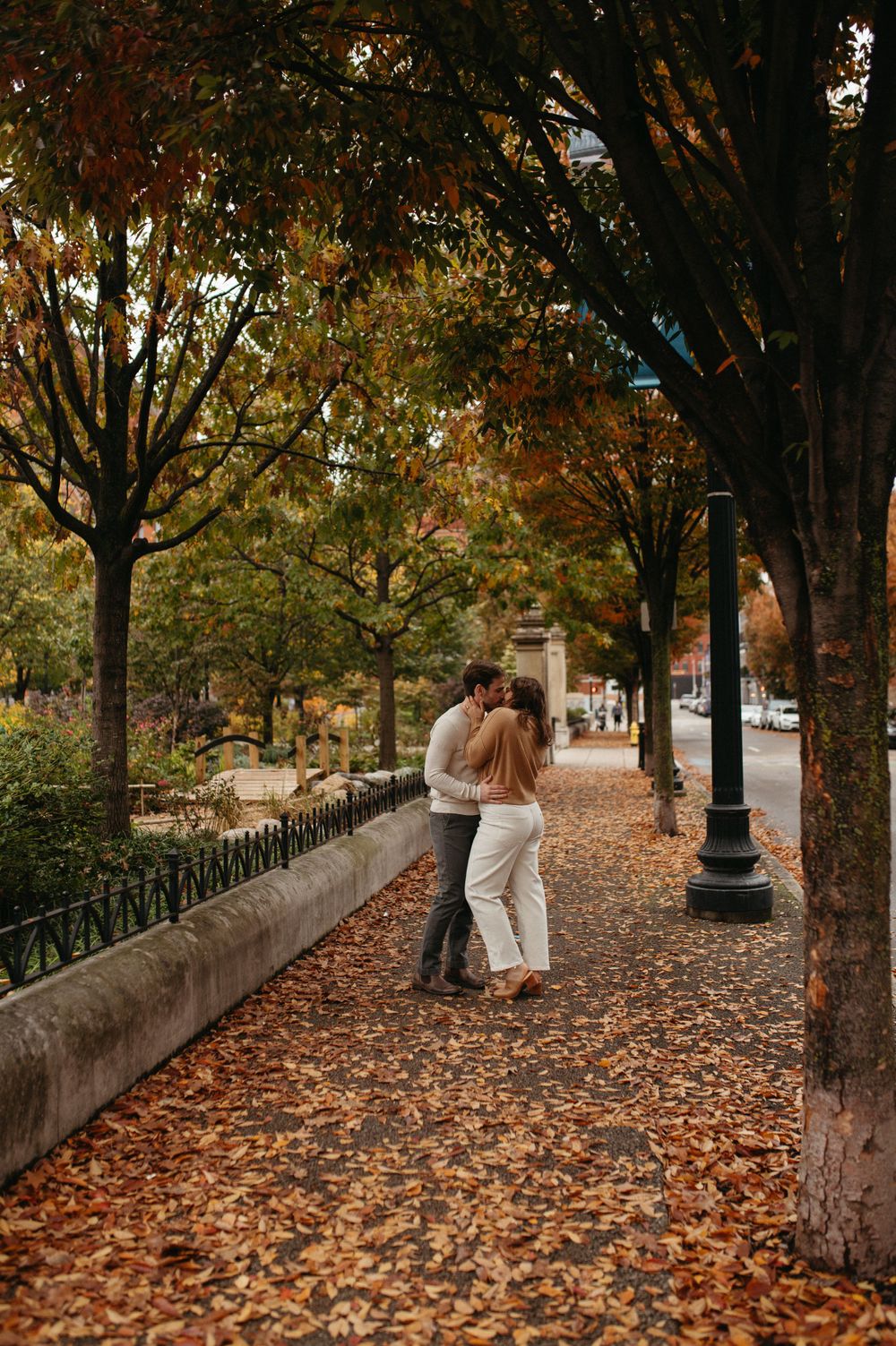 A couple sharing an intimate moment on a tree-lined path covered in fallen autumn leaves.