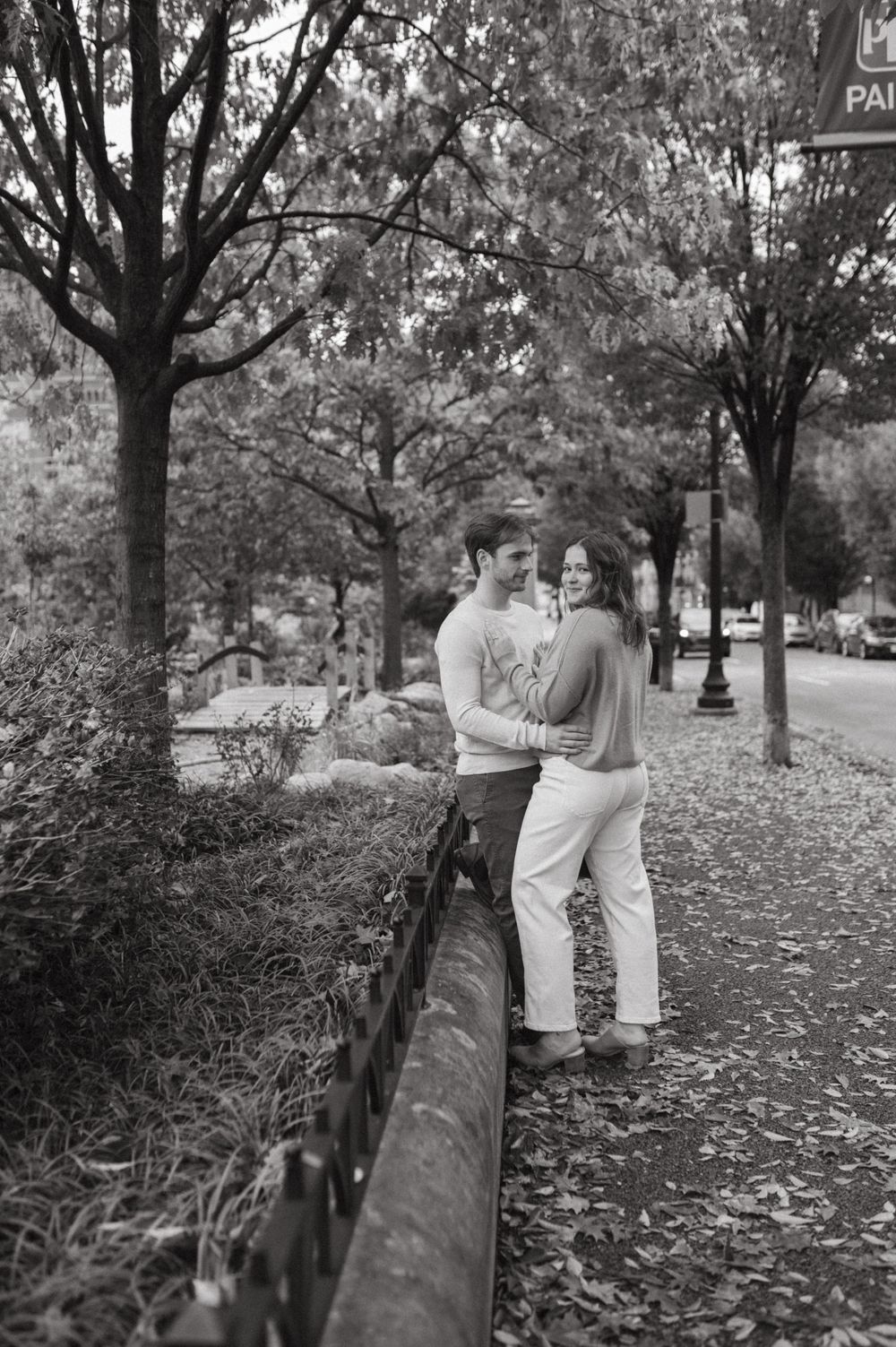 Black and white photo of couple by wrought iron fence on leaf-covered path.