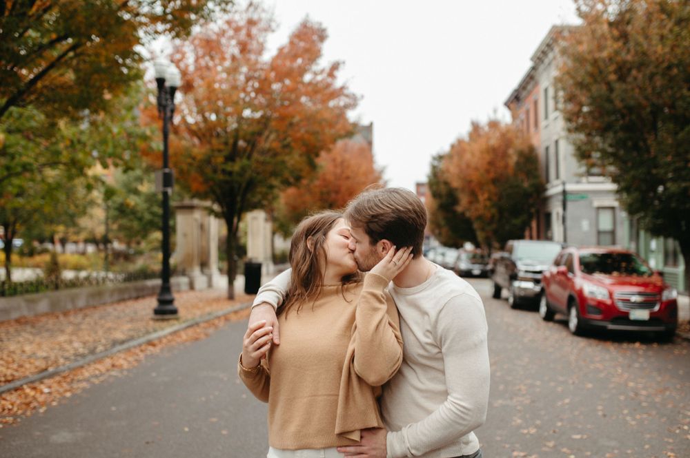 Couple embracing on tree-lined street with autumn colors and parked cars.