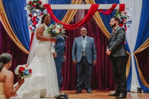 Wedding ceremony under red and blue draped backdrop with floral arrangements.