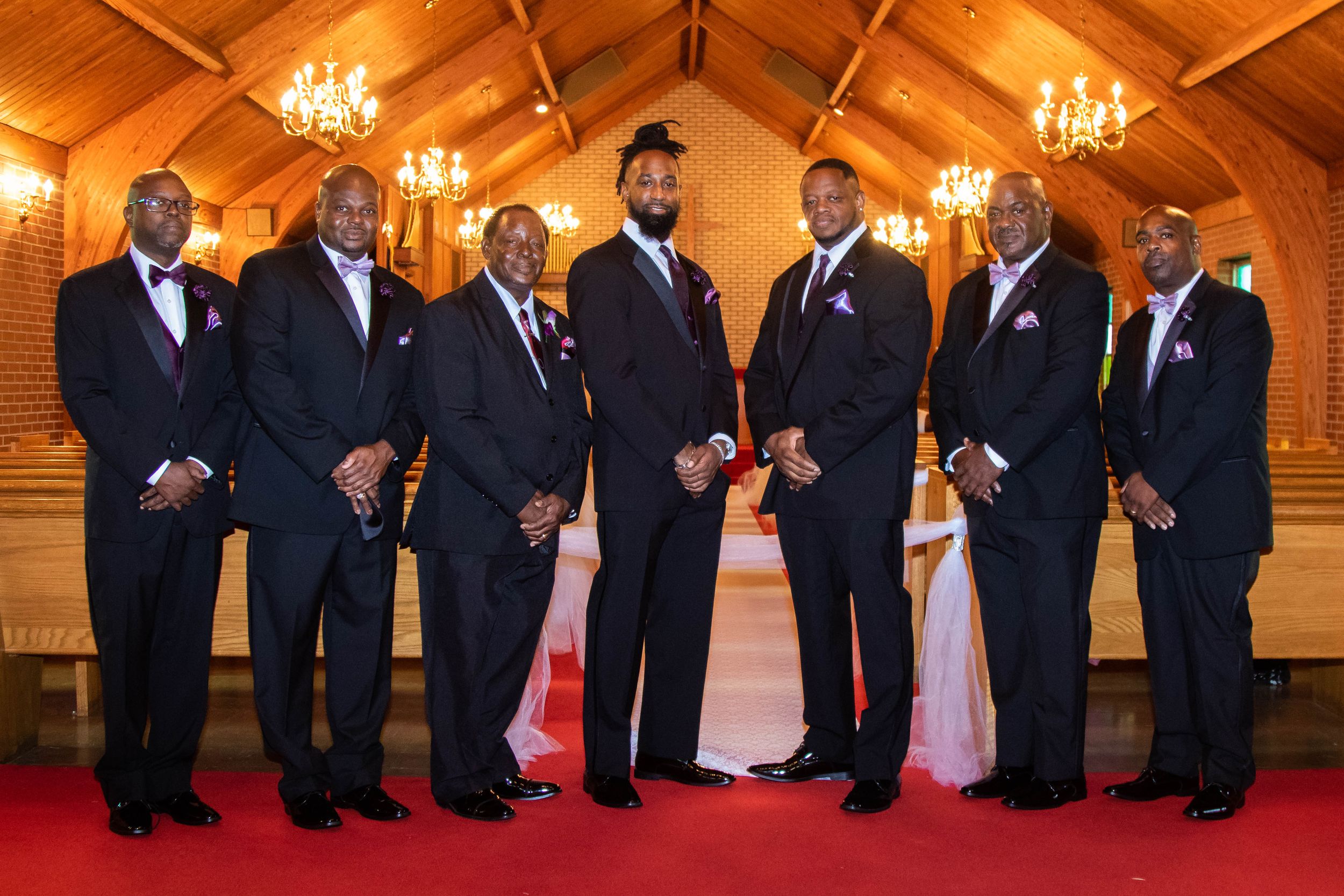 Wedding party in black suits poses formally in wooden chapel with chandeliers.
