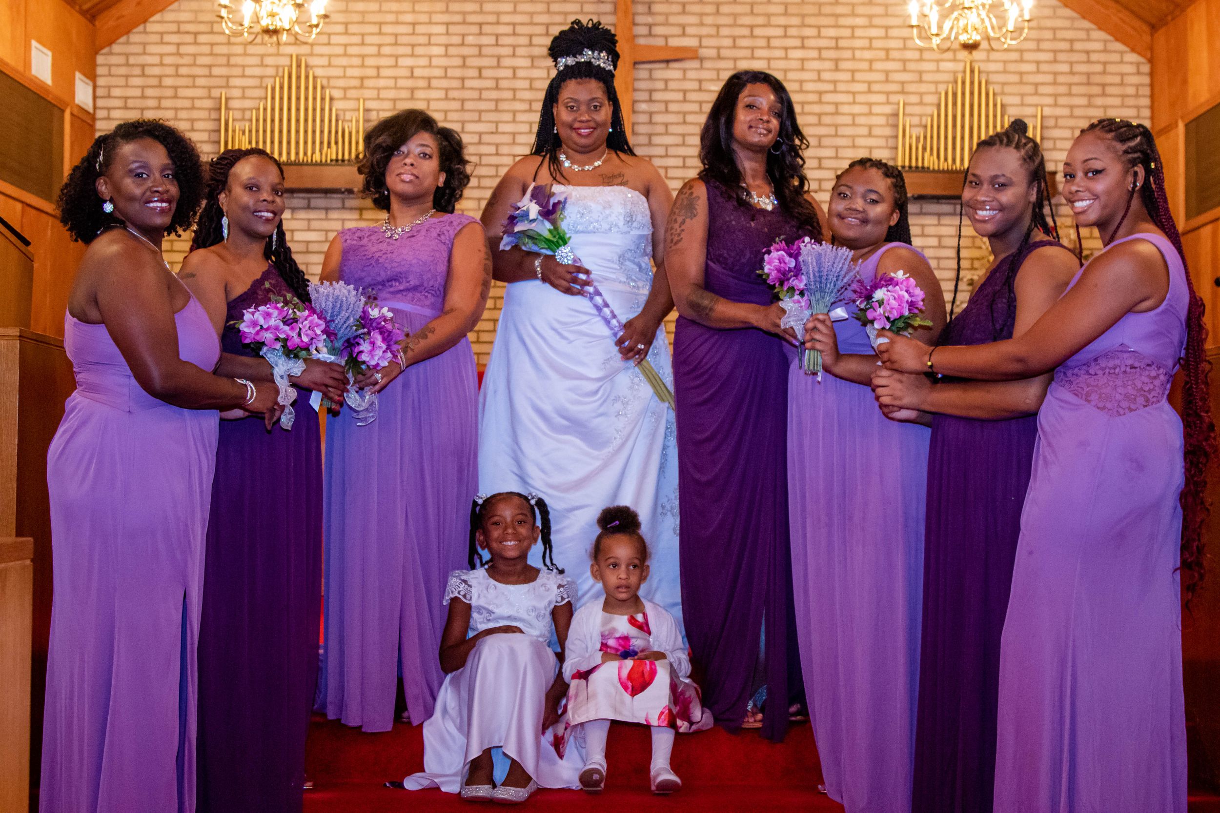 Bridal party in purple dresses and white gown poses together with flower girls on red carpet.