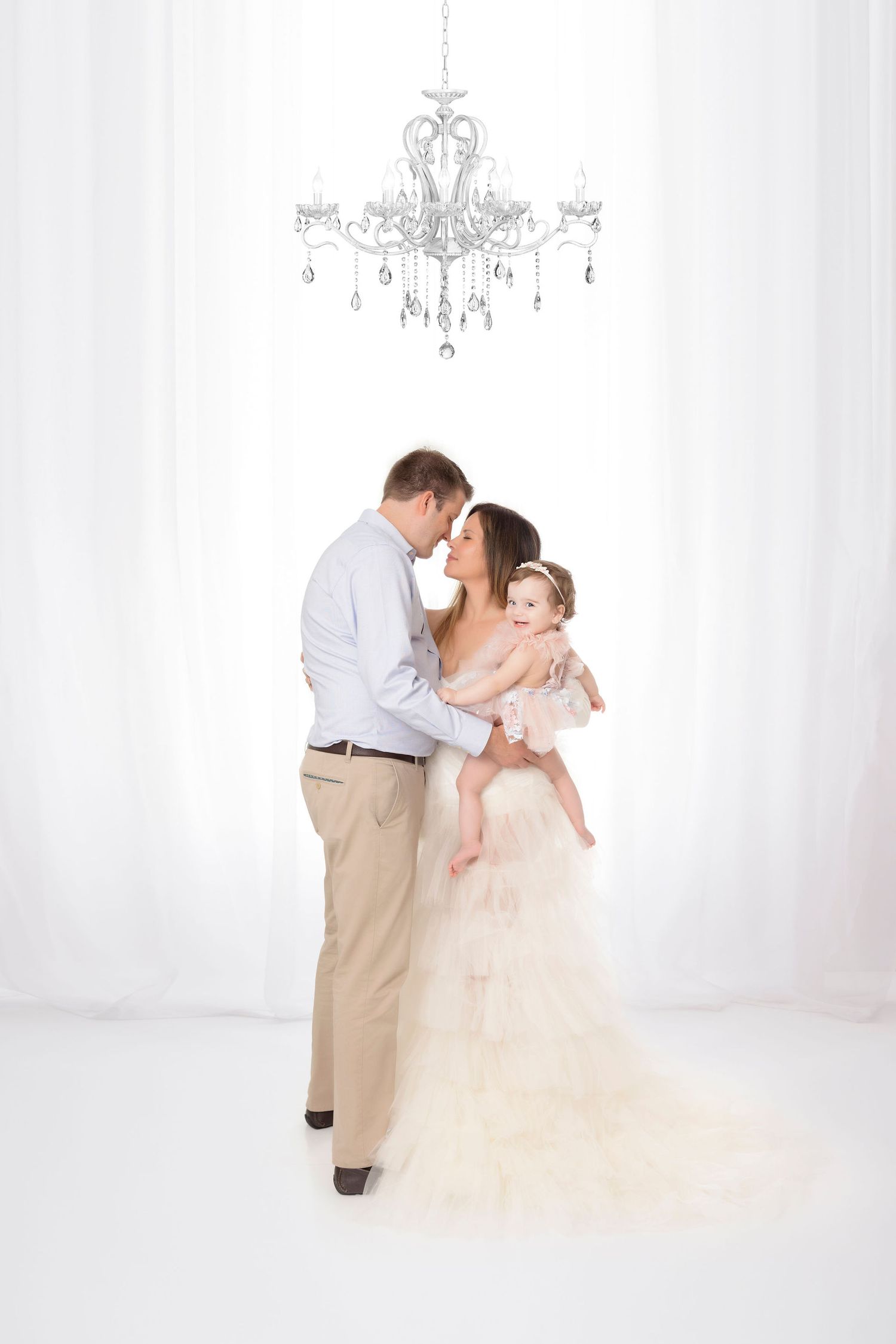 Intimate family moment under crystal chandelier in elegant white studio setting.