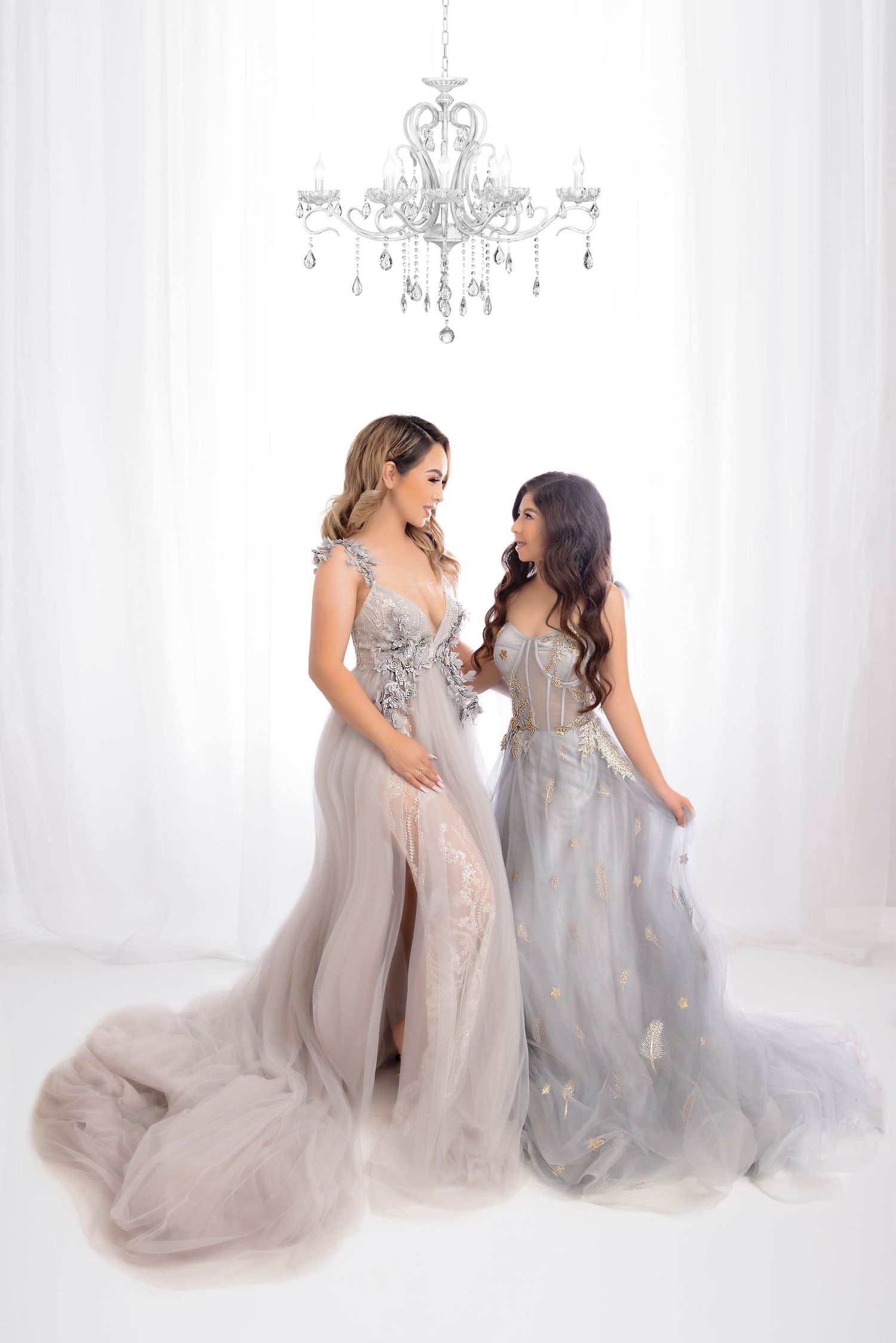 Two women in stunning gray formal gowns pose beneath crystal chandelier.