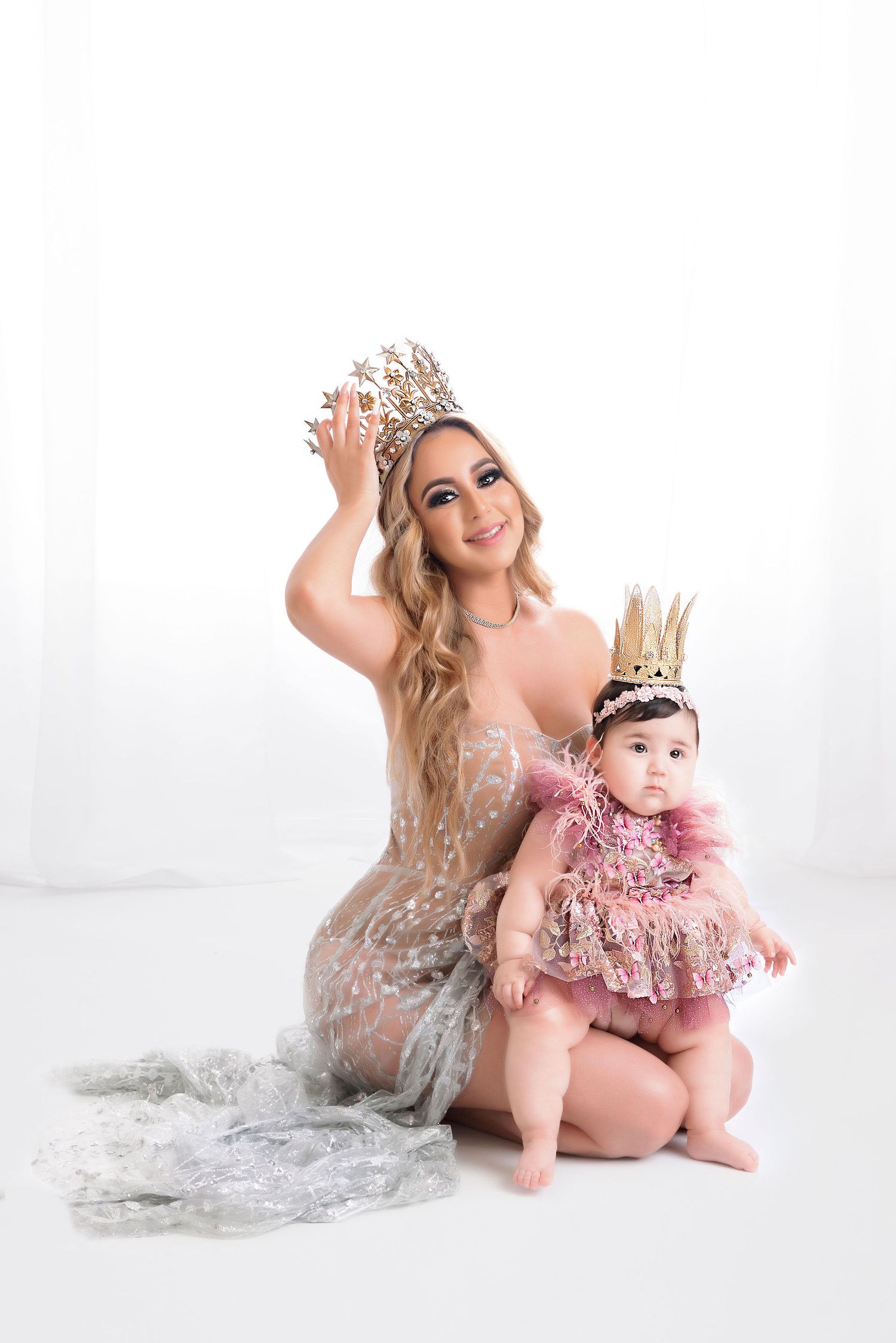 Two figures in matching crowns share a tender moment wearing glamorous dresses against a bright white background.