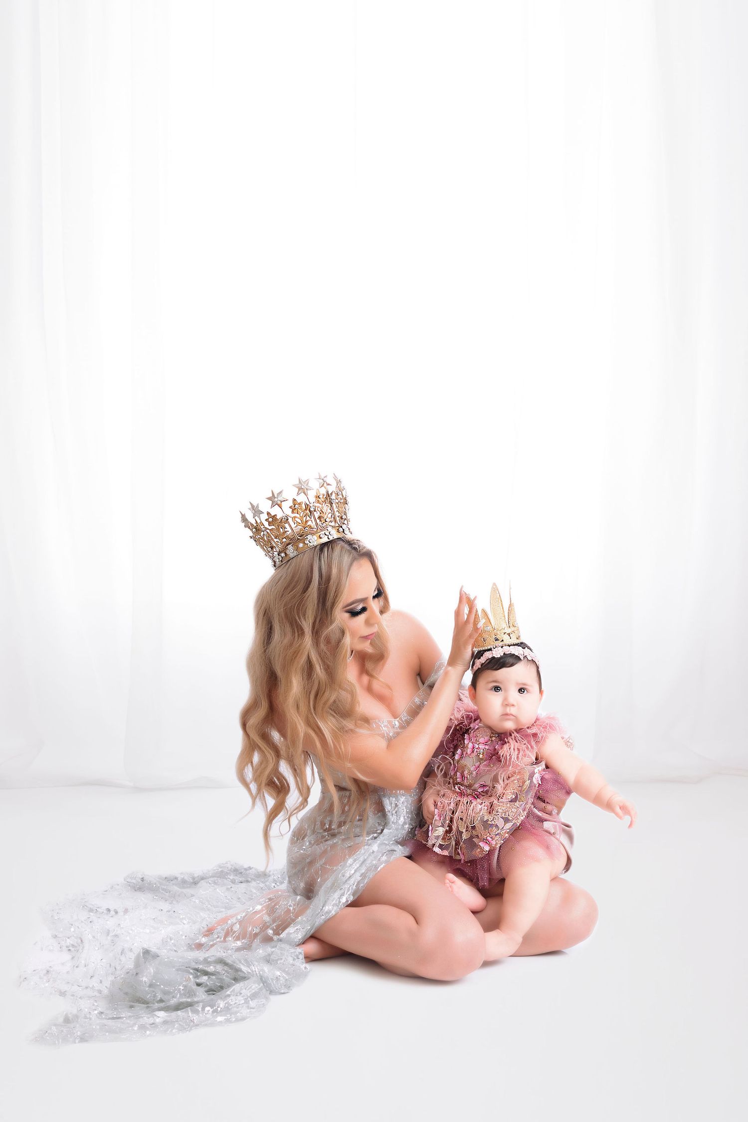 A woman in a silver dress and crown holds a baby wearing a pink dress and matching crown against a white backdrop.