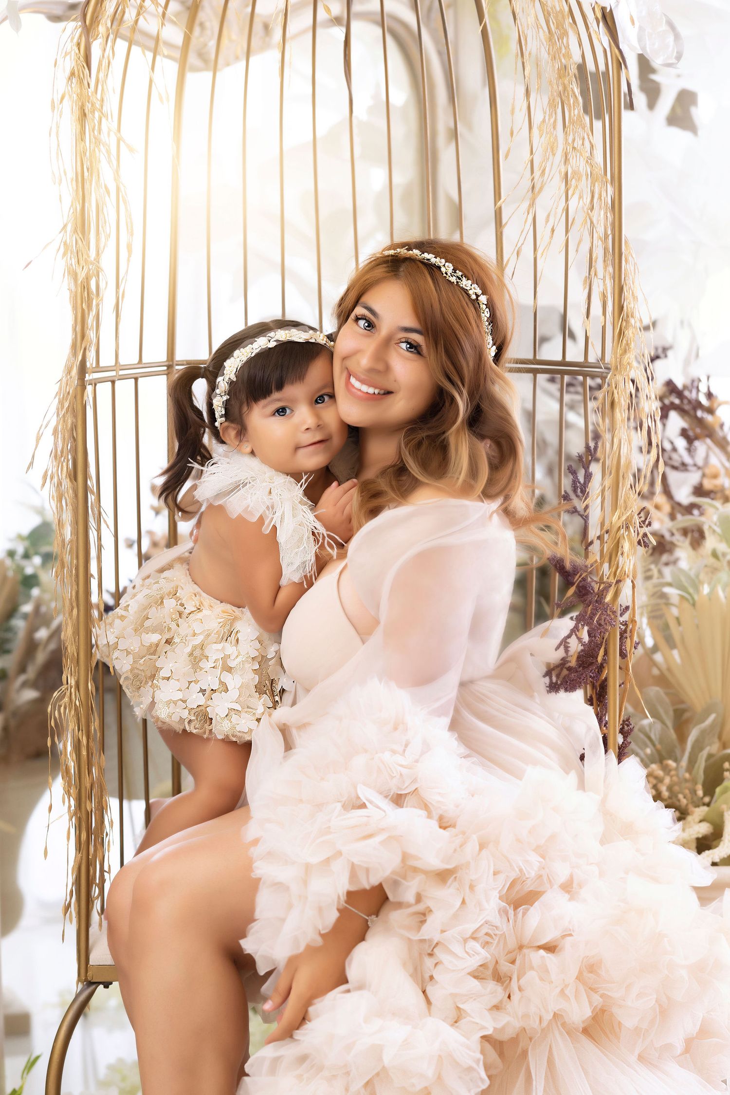 An elegant portrait featuring two people in light dresses posing by a decorative gold birdcage.