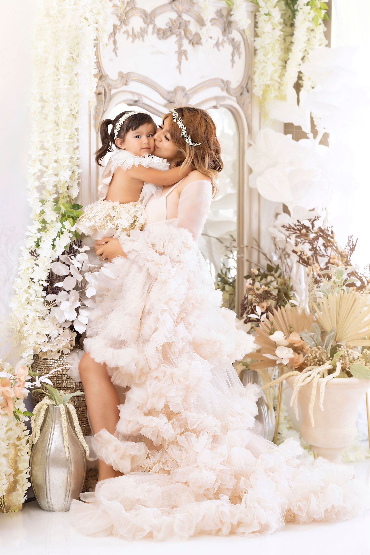 Two figures in flowing white dresses embrace among floral decorations and dried pampas grass.