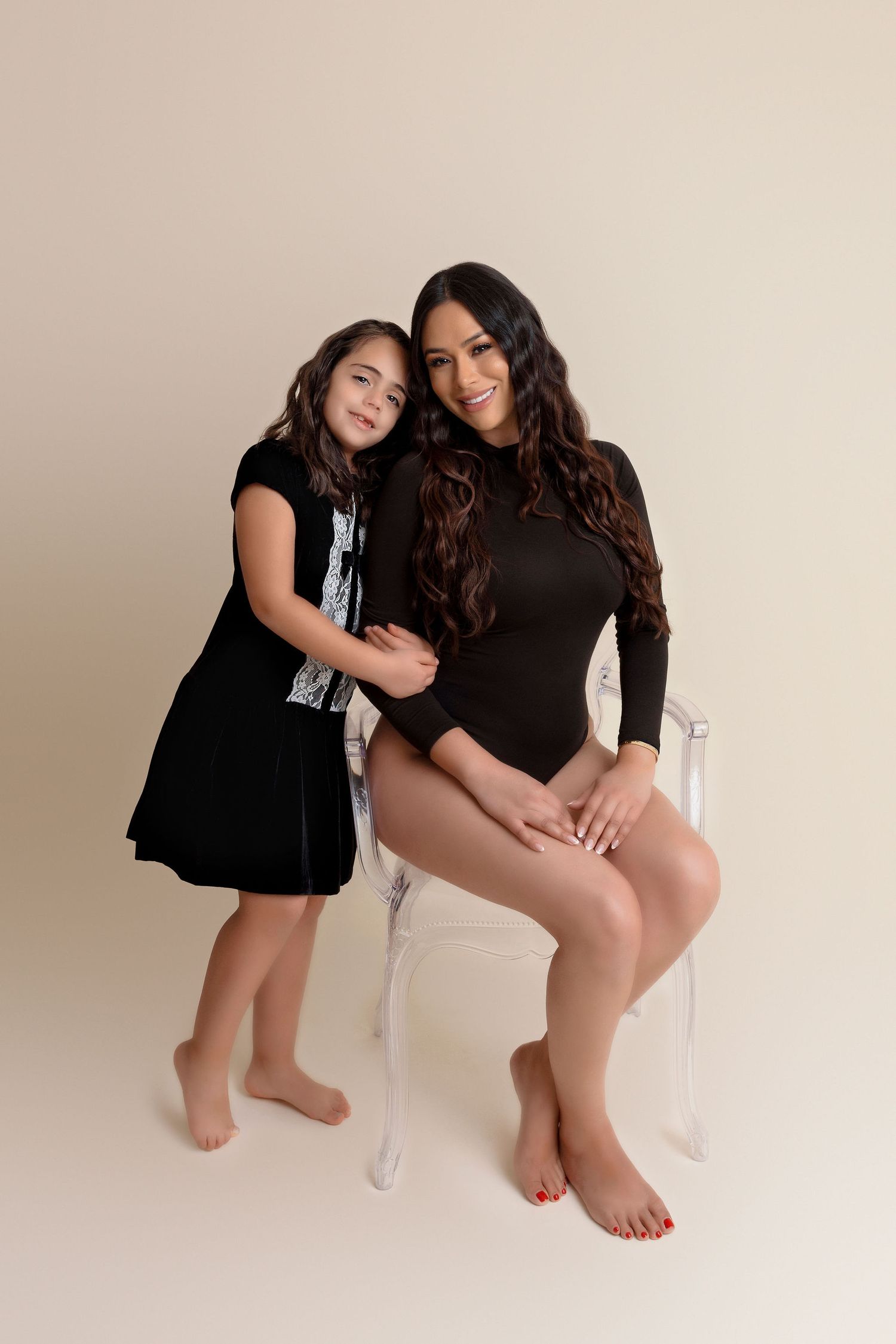 Two people in black outfits pose together on a clear acrylic chair against a neutral backdrop.