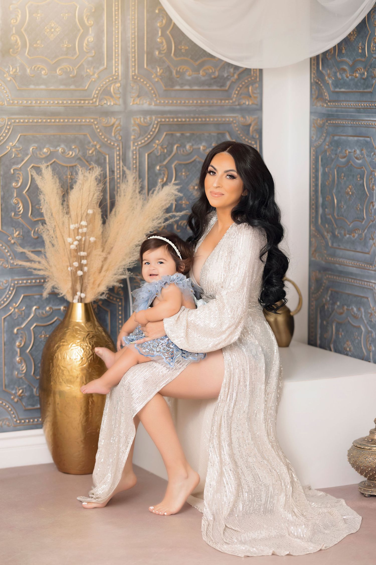 Elegant portrait session featuring flowing white dress and gold accents against ornate blue wall with decorative vase.