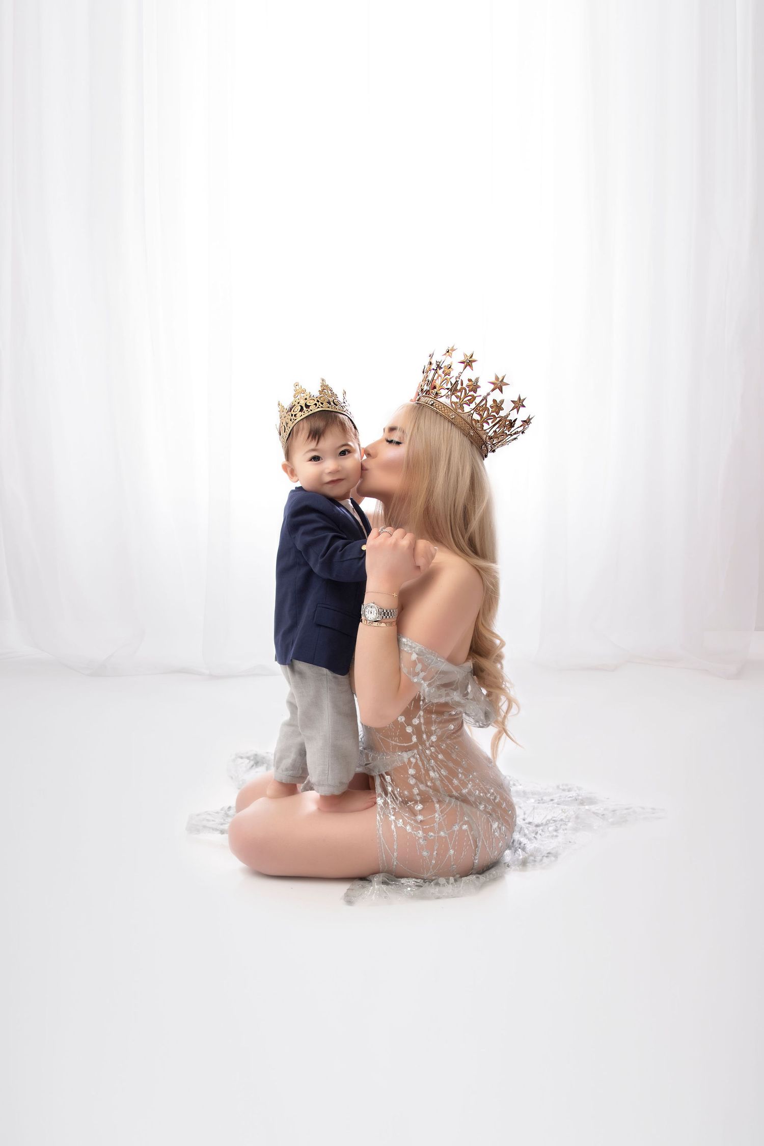 Elegant mother and child in formal attire wearing crowns pose together against white background.