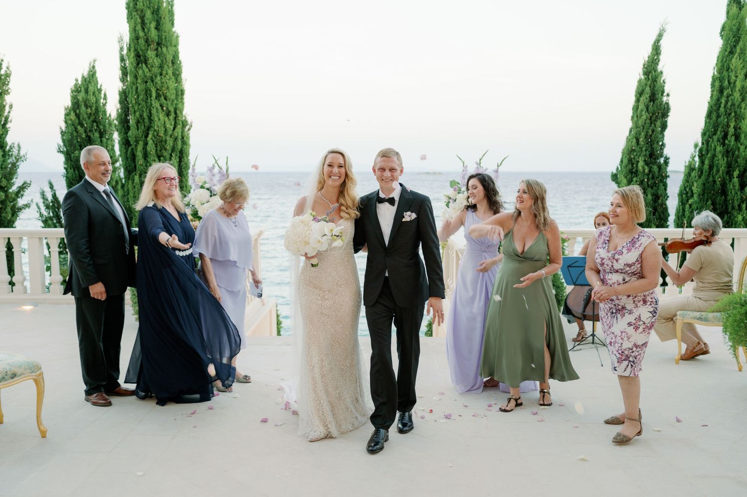 Elegant wedding party poses on a seaside terrace with cypress trees and ocean views.