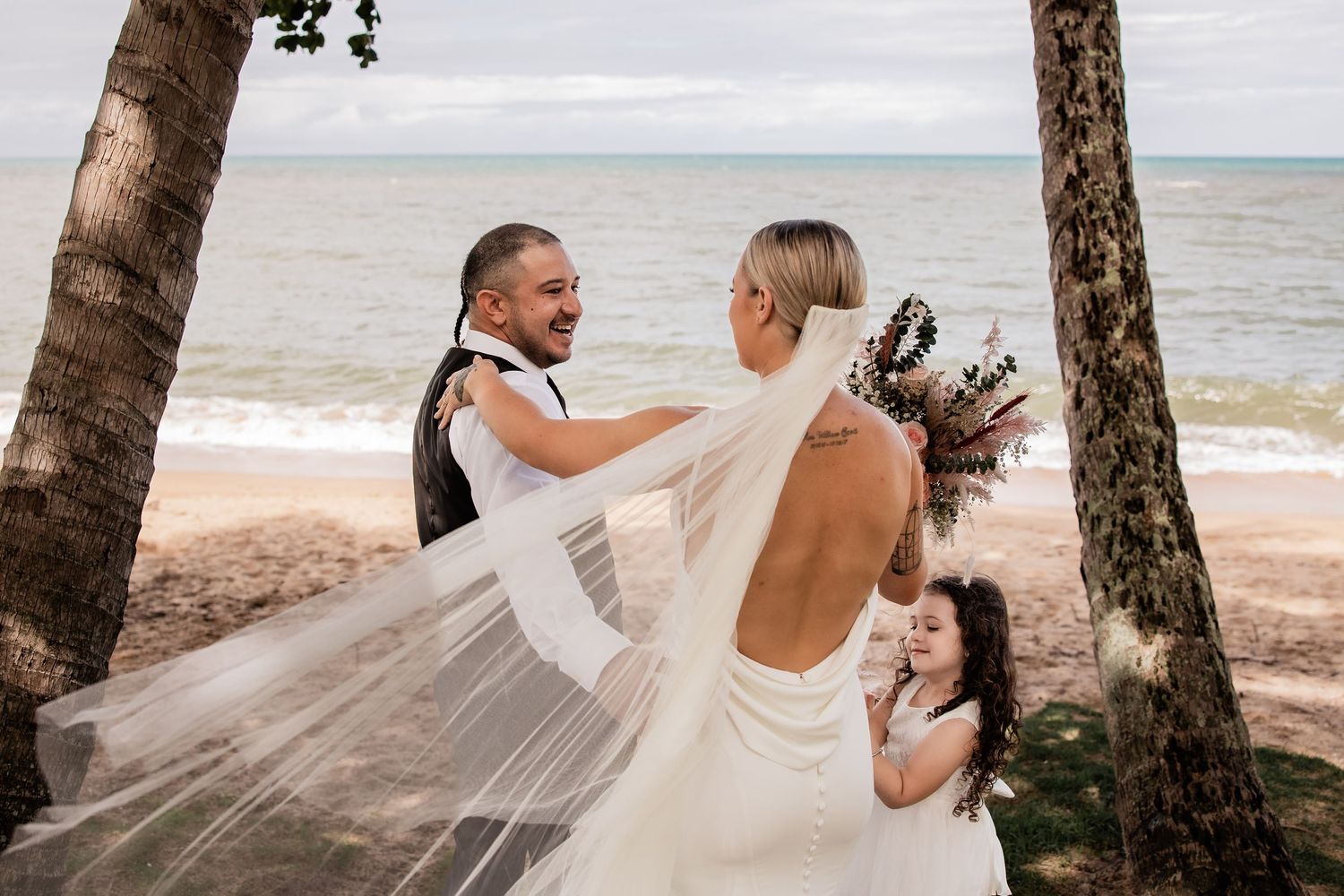 Couple shares romantic moment on beach with palm trees as bride's veil flows in wind.