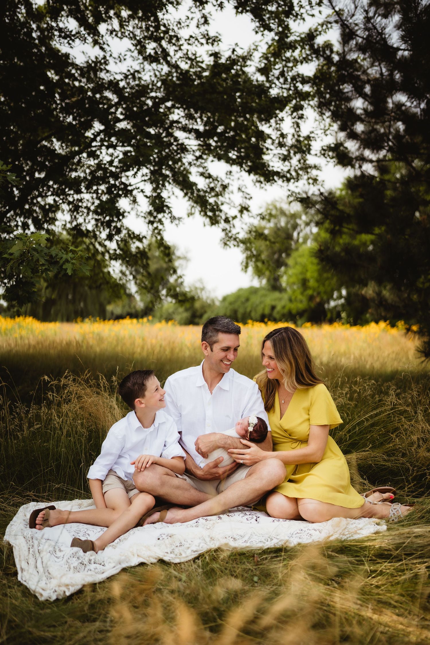 Family sits together on white blanket in golden sunlit meadow during outdoor photoshoot.