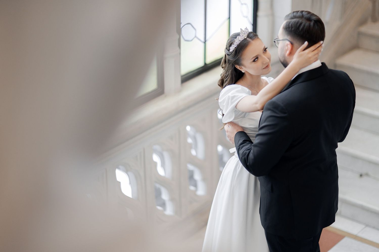 A romantic wedding moment as couple embraces on grand staircase with natural light streaming through windows.
