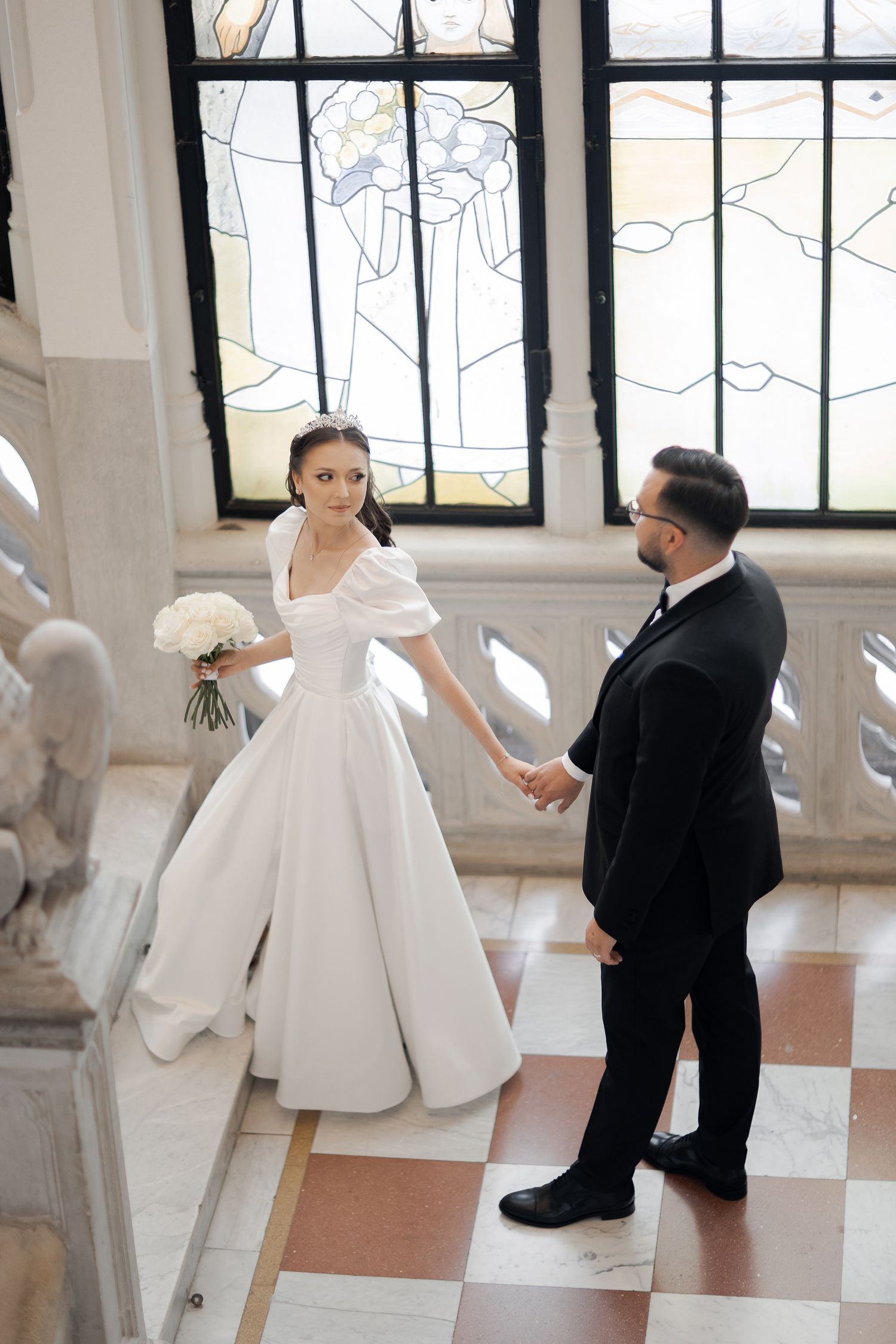 Elegant couple in formal wedding attire pose in bright interior with stained glass windows.