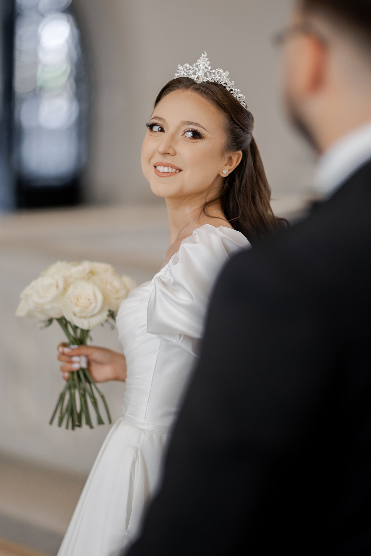 Bride in classic white gown and tiara holds white bouquet while smiling over her shoulder.
