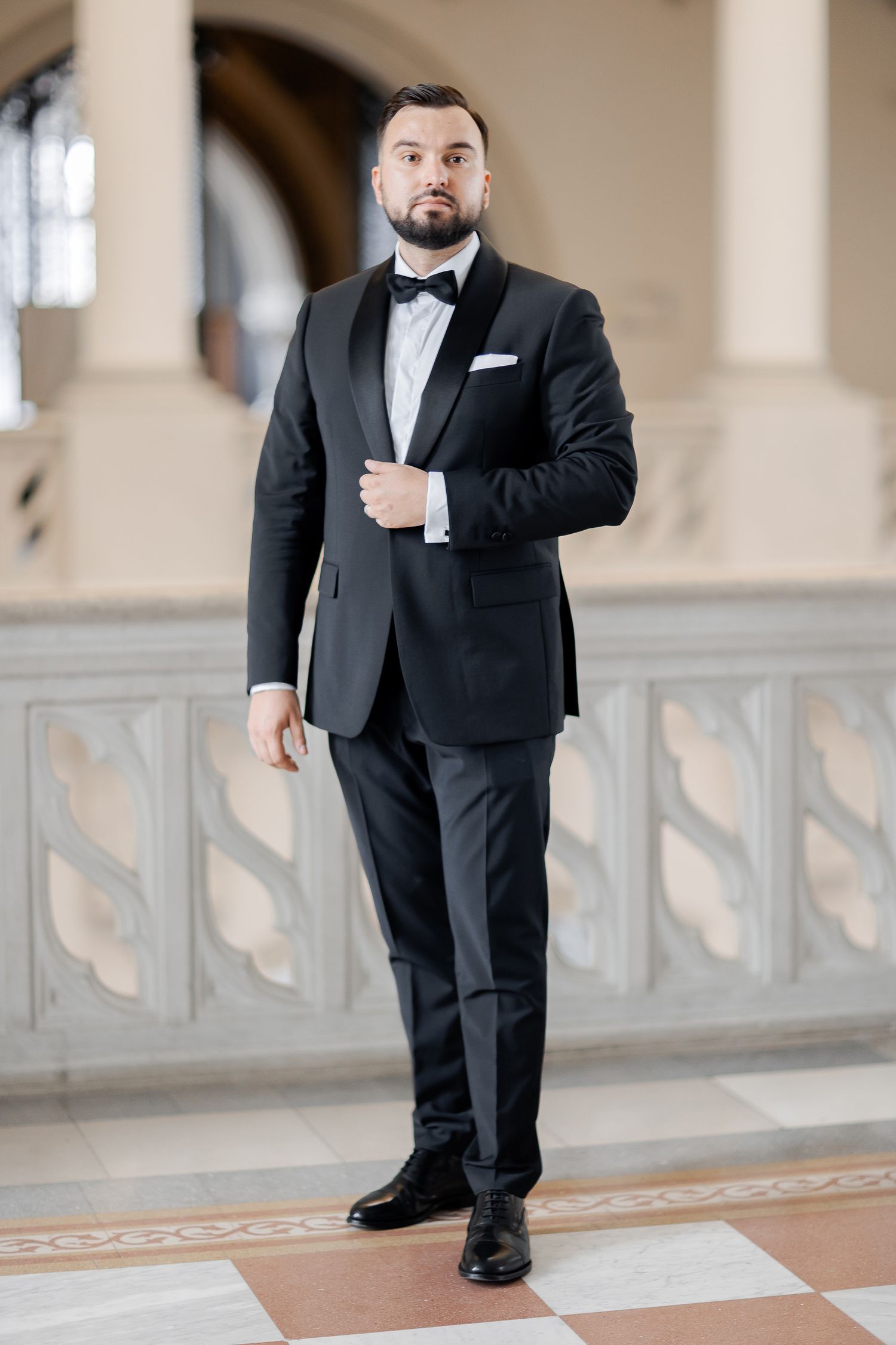 Groom in classic black tuxedo and bow tie stands confidently in elegant hallway setting.