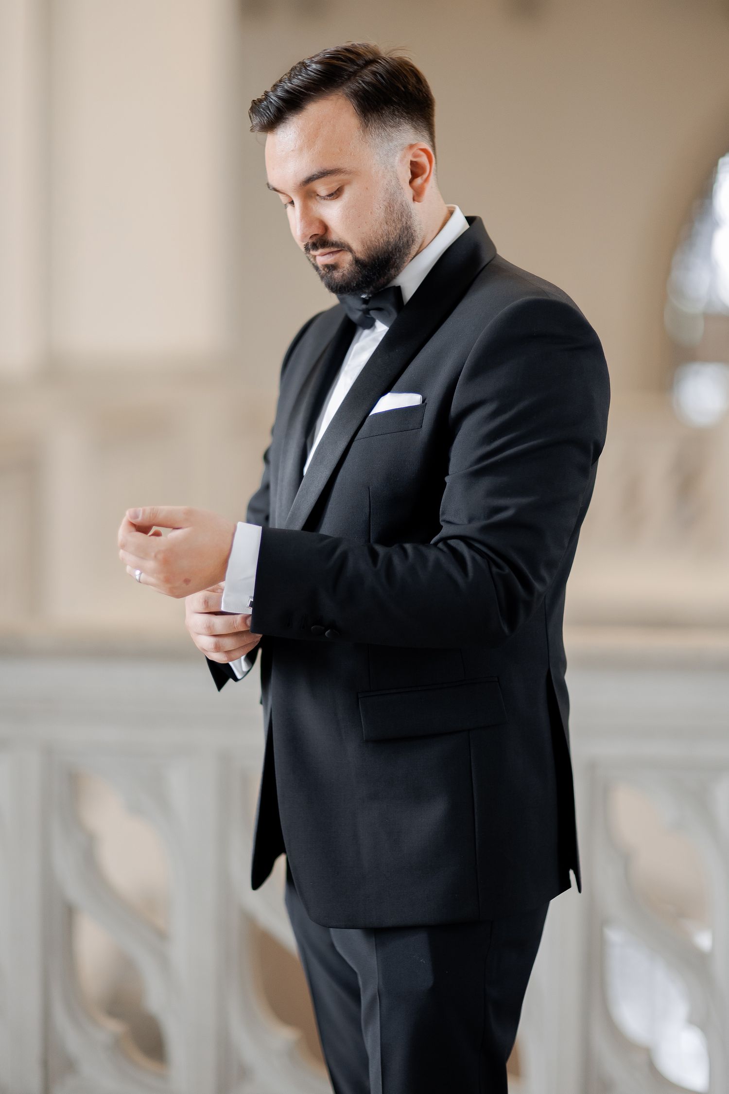 Groom adjusts cufflink while wearing formal black tuxedo in classical architectural setting.