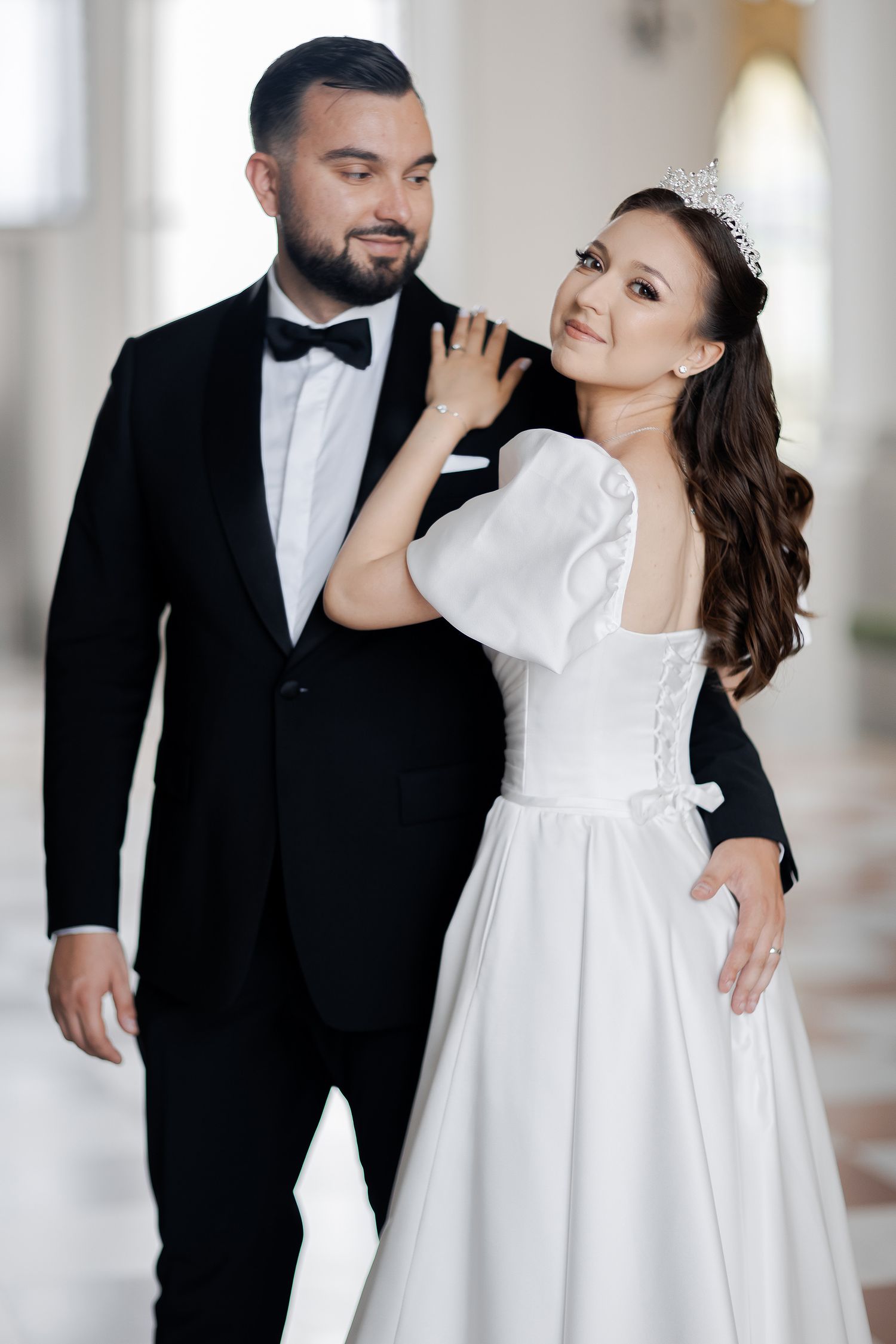 Elegant couple in formal wedding attire pose together in bright hallway with classical architecture.
