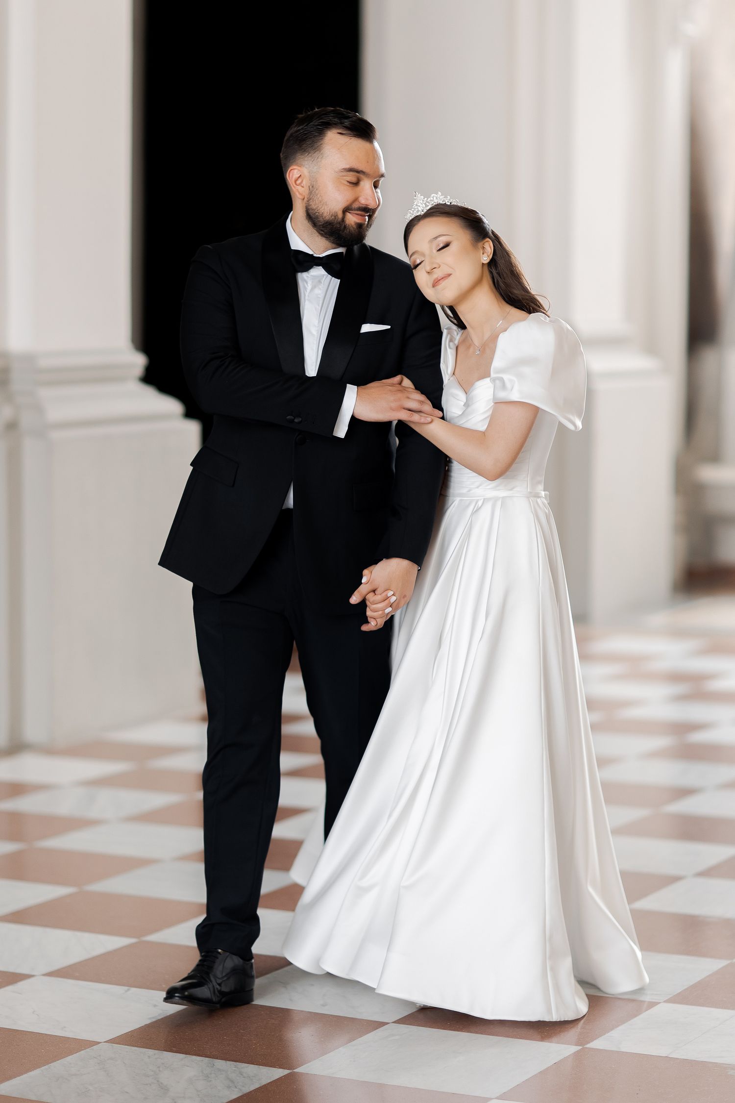 Wedding couple dancing together on checkerboard floor in classical white hallway.