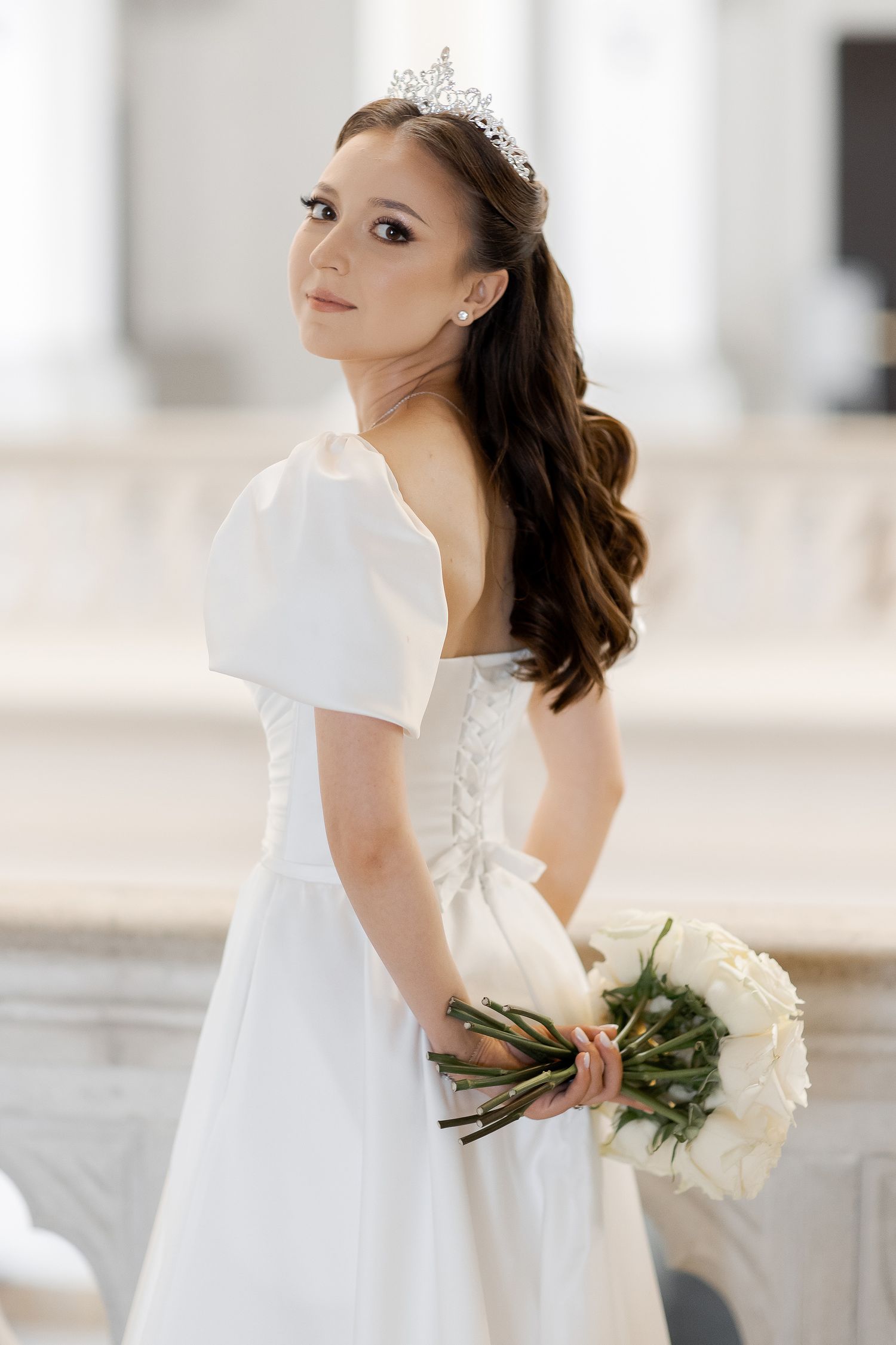A bride in a white wedding gown with puffy sleeves and tiara poses with white roses.