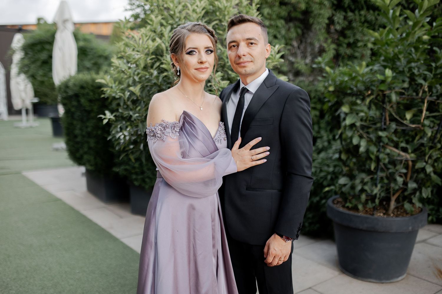 A couple poses outdoors in formal attire, with her wearing a lovely lavender dress.