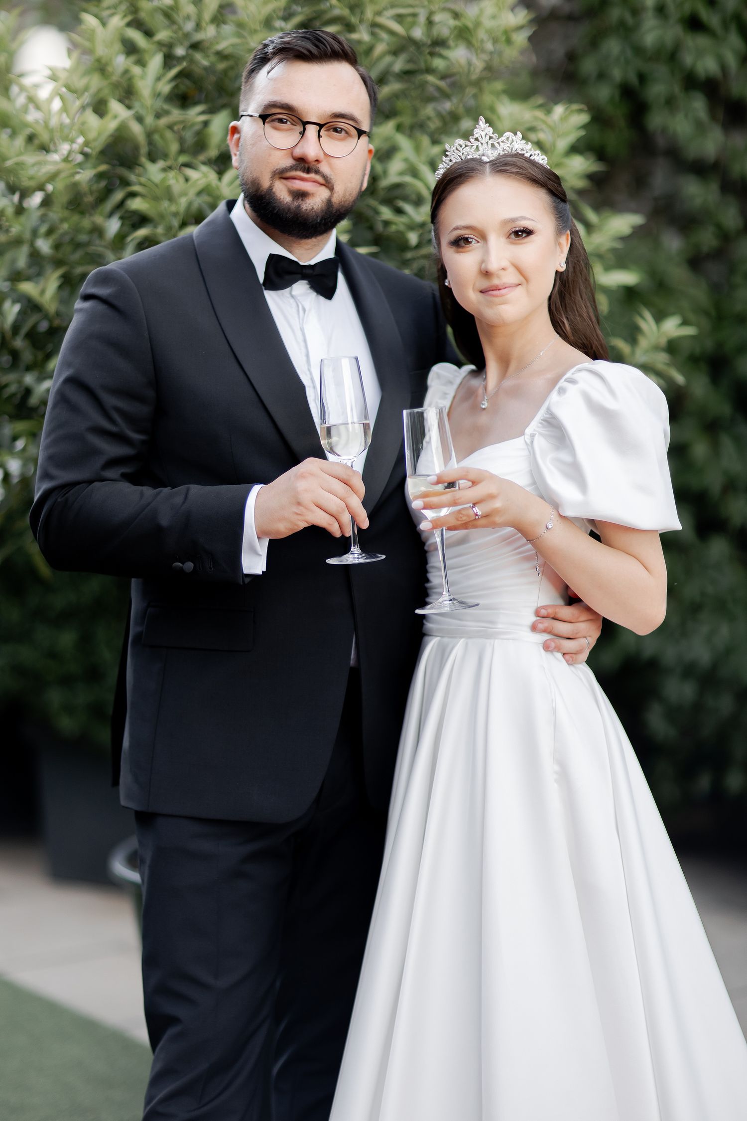 A wedding couple toasts with champagne glasses in formal attire against a garden backdrop.