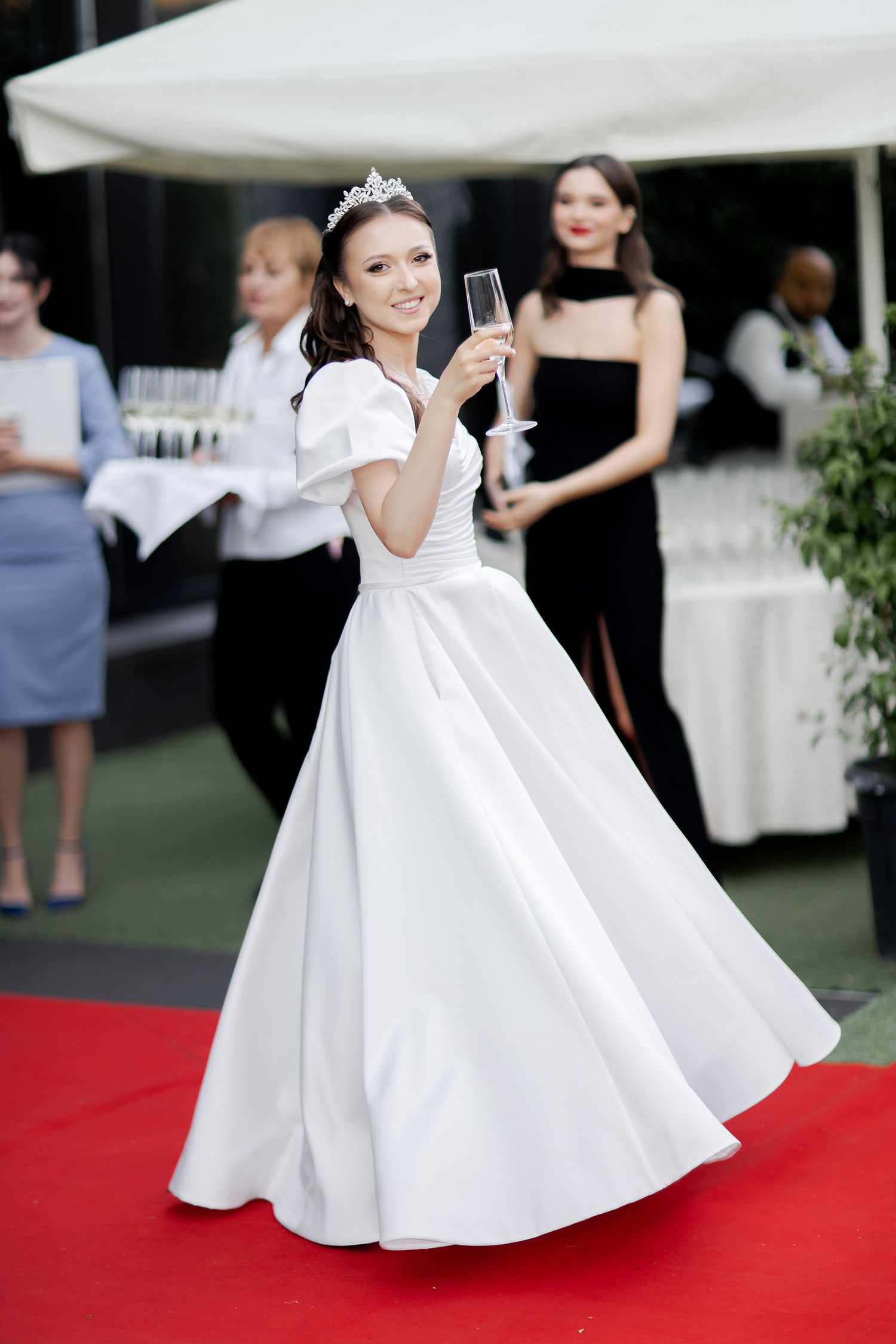 Elegant woman in flowing white wedding dress and tiara poses with champagne glass on red carpet at outdoor reception.