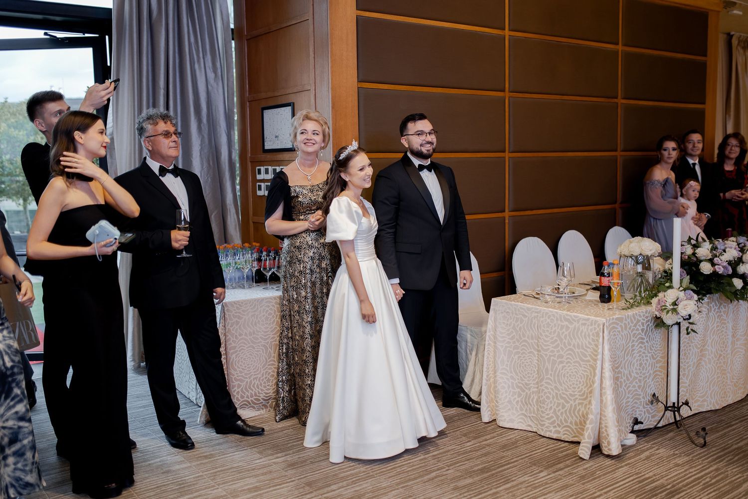 Wedding reception guests in formal attire gather in elegant venue with wood paneled walls.