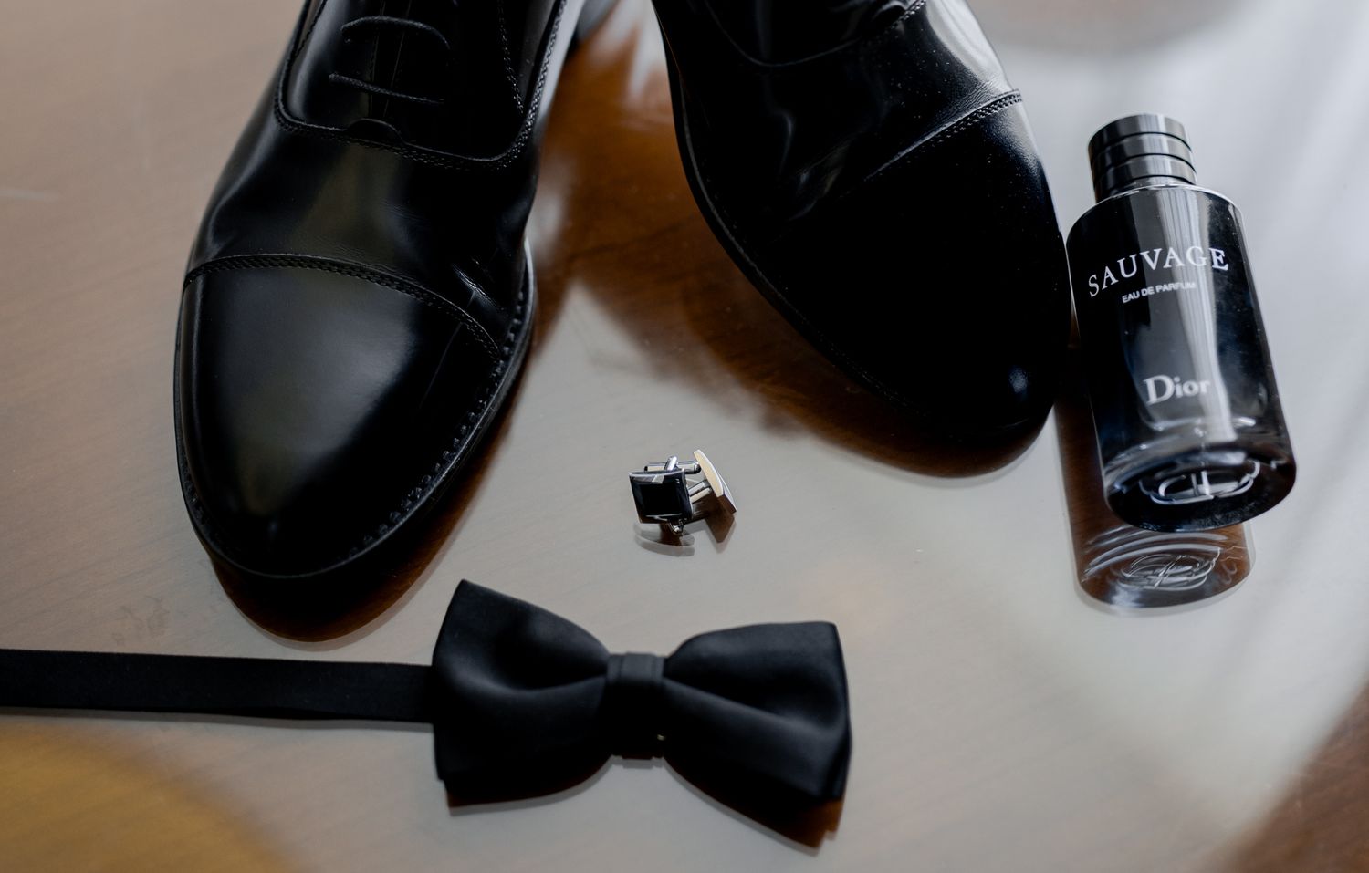 Wedding accessories including black dress shoes, bow tie, and cologne bottle arranged on a surface.