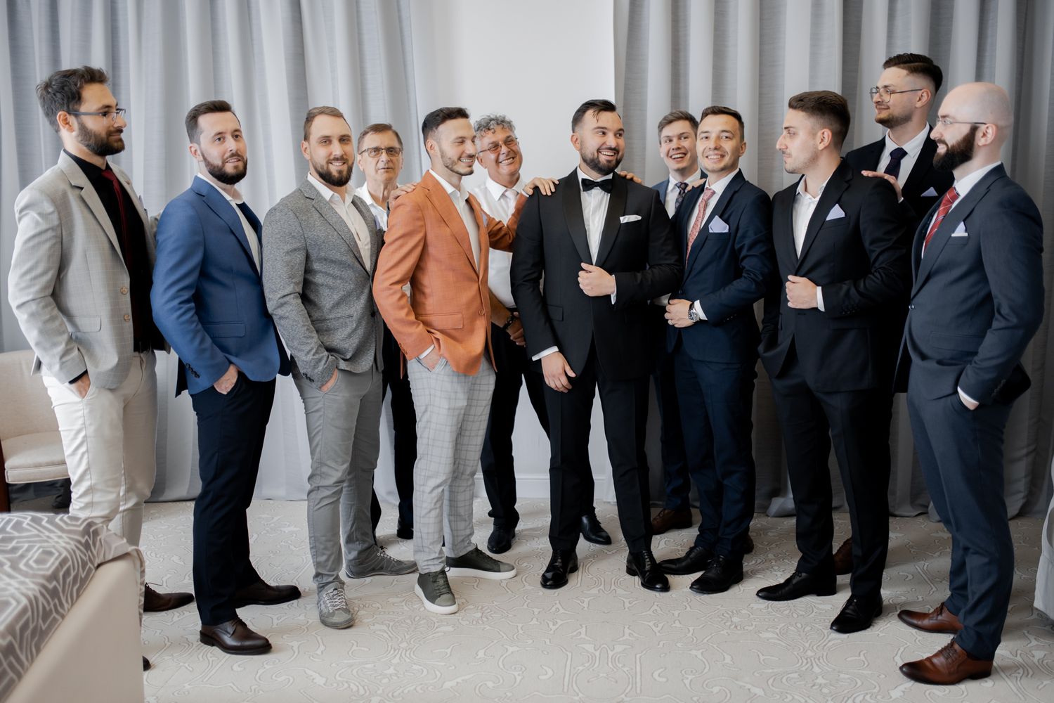 Wedding party in coordinated suits ranging from light to dark colors pose together indoors.
