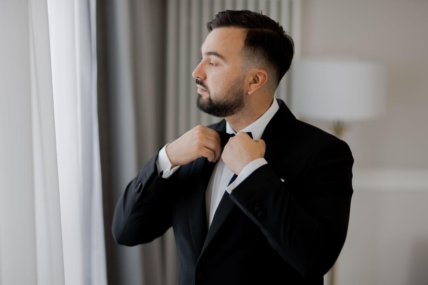 Person in black tuxedo adjusting bow tie near window with natural light.