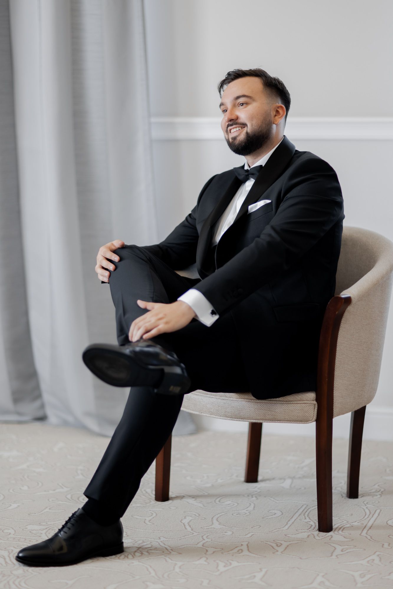 Person in classic black tuxedo seated elegantly in beige armchair.