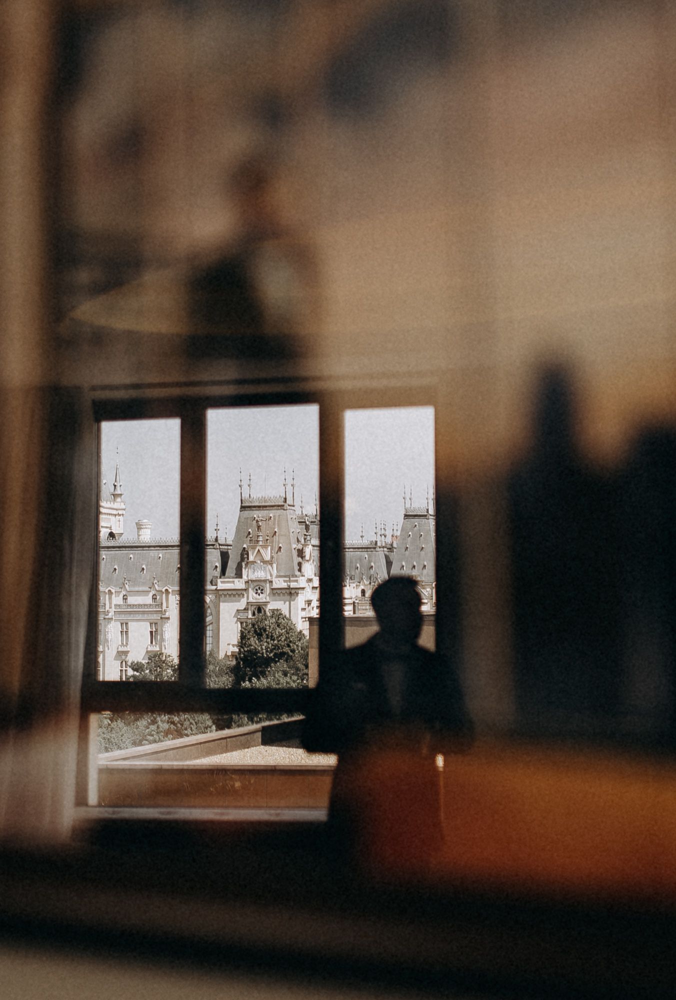 Silhouetted figure gazes through window at historic European architecture and spires in moody atmospheric scene.
