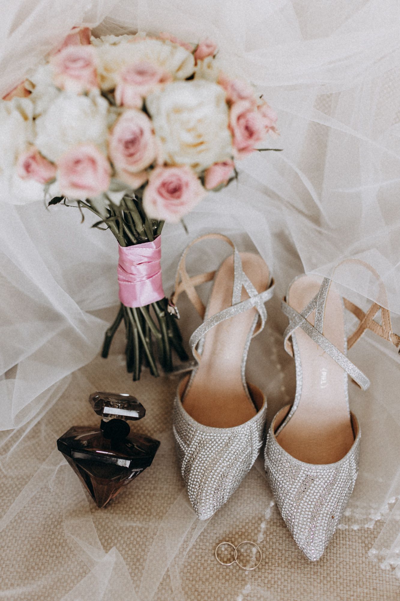 Pink and white rose bouquet with sparkly bridal shoes and perfume bottle on delicate tulle fabric.